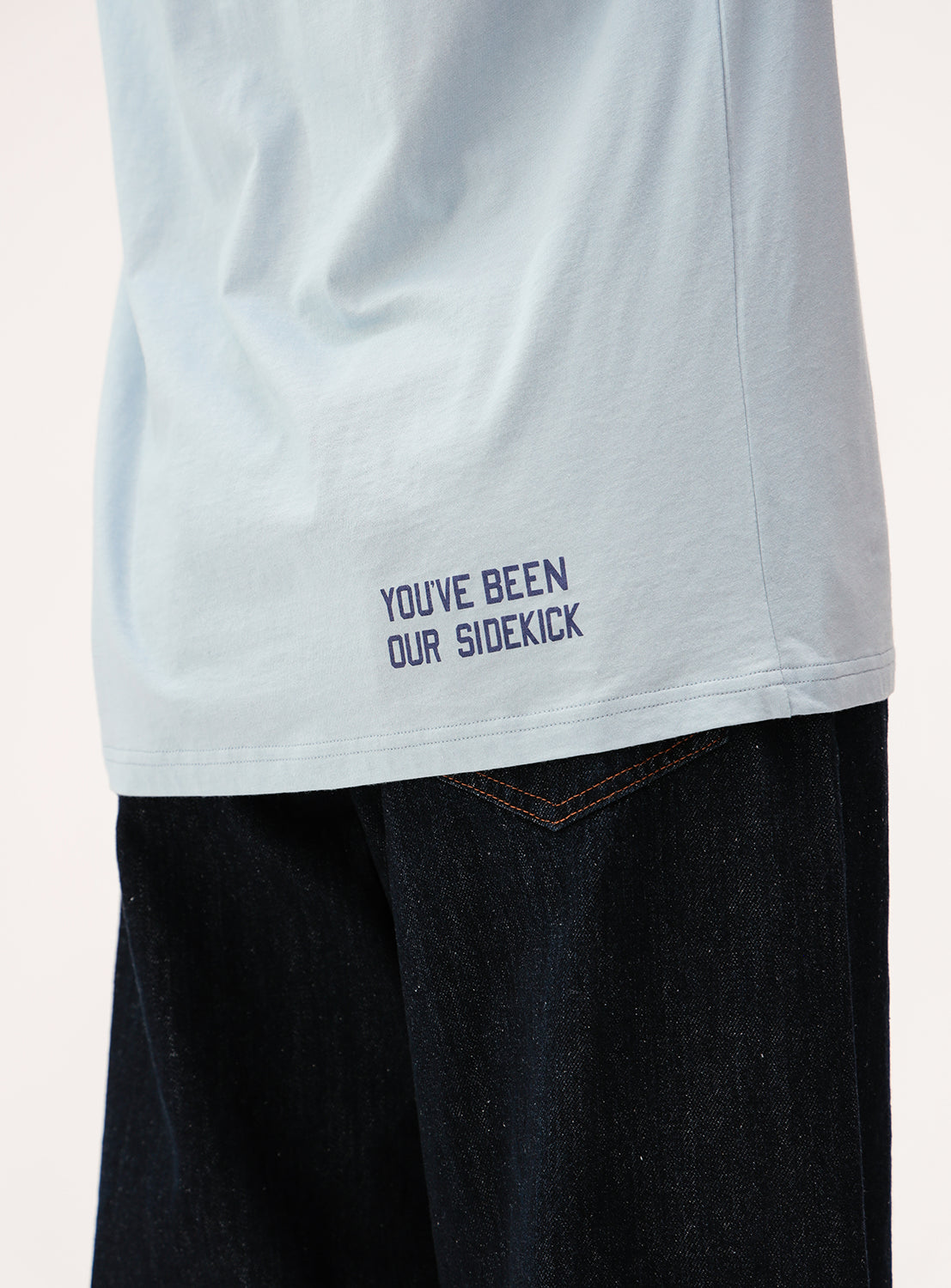 A person wearing the BEAMS BOY Big Logo Long-Sleeve T-Shirt in light blue, featuring “YOU’VE BEEN OUR SIDEKICK” stitched near the hem, paired with dark jeans.