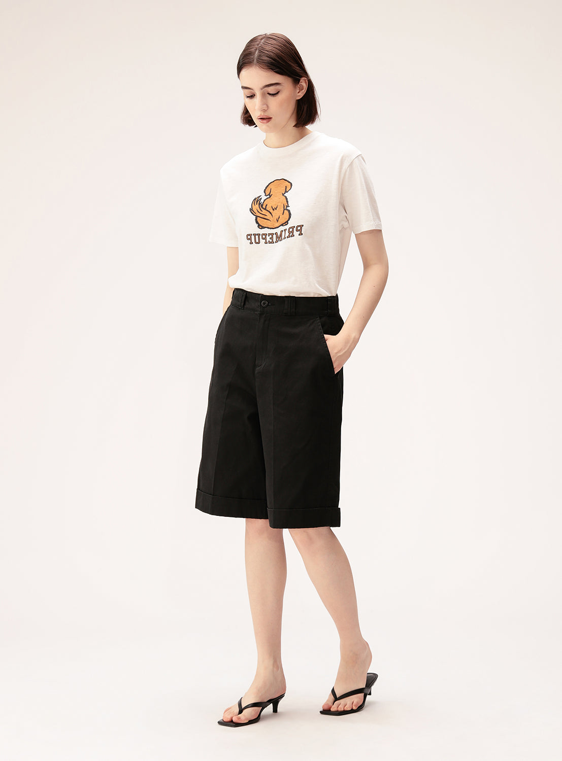 A woman stands with her hands in her pockets, wearing the BEAMS BOY Preppy Dog Graphic T-Shirt, black knee-length shorts, and black sandals against a plain light background.