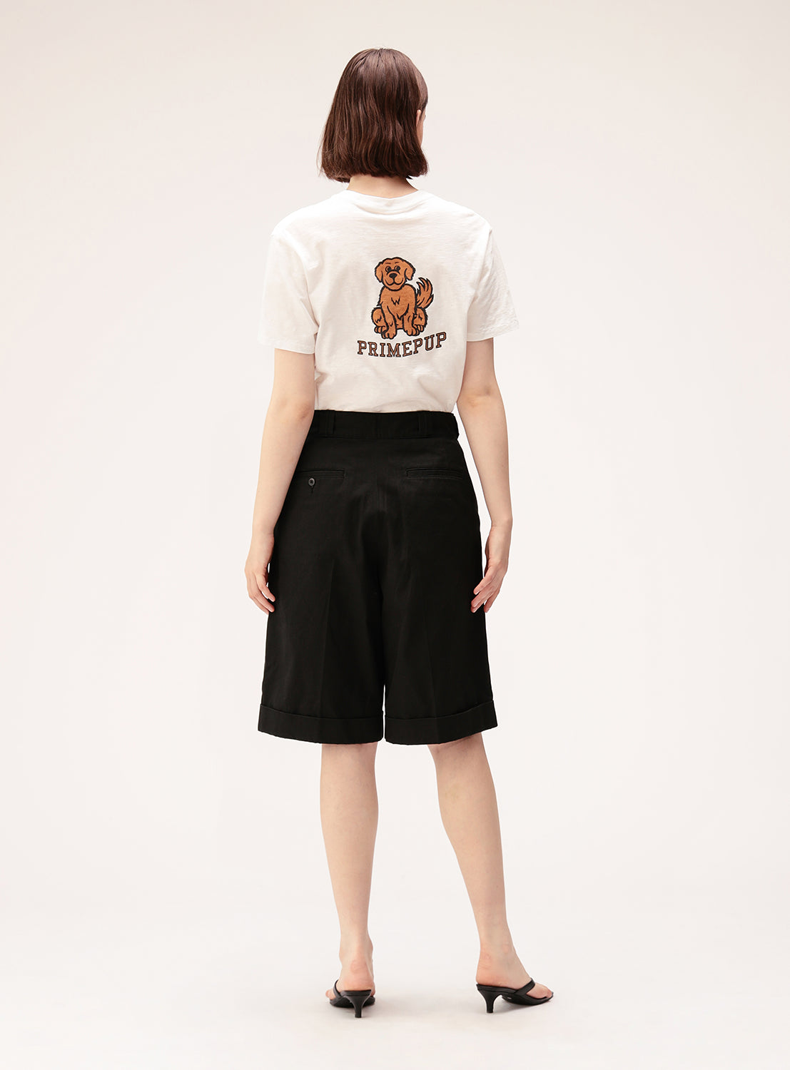 A person facing away wears the BEAMS BOY Preppy Dog Graphic T-Shirt, styled with black knee-length shorts and black heeled sandals for a subtle preppy look. The background is plain and light-colored.
