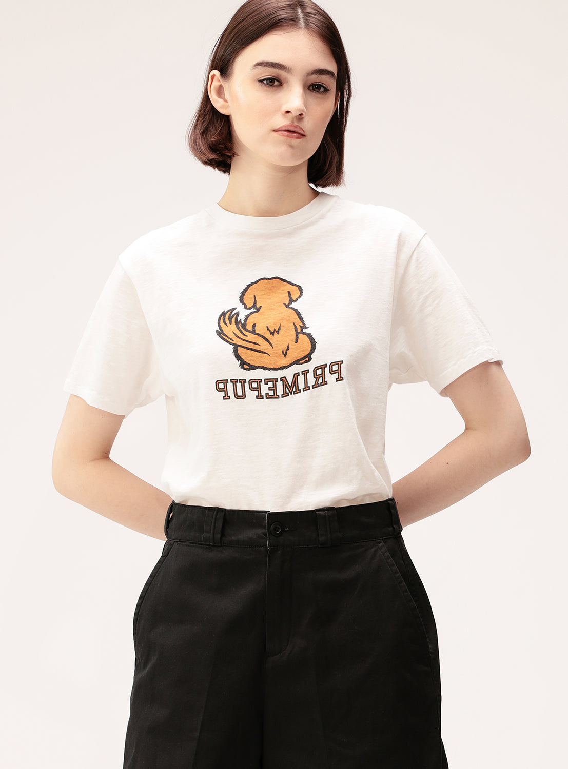 A woman with short brown hair wears the BEAMS BOY Preppy Dog Graphic T-Shirt, a vintage-inspired cotton tee featuring an illustrated dog and bold "PRIMEPUP" text, styled effortlessly with high-waisted black pants for a cool preppy look.