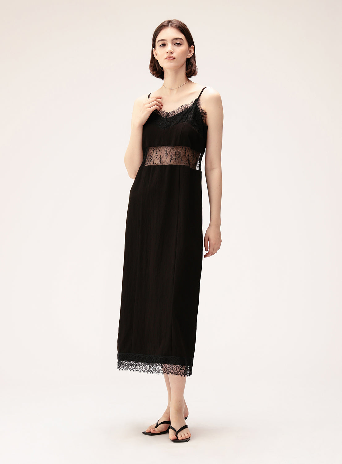 A woman with short brown hair stands against a plain light background wearing the Ray BEAMS Lace Cut-Out Cami Dress, featuring delicate lace trim at the neckline, waist, and hem. Styled with black sandals and a neutral expression, this lingerie-inspired dress lends her an understated elegance.