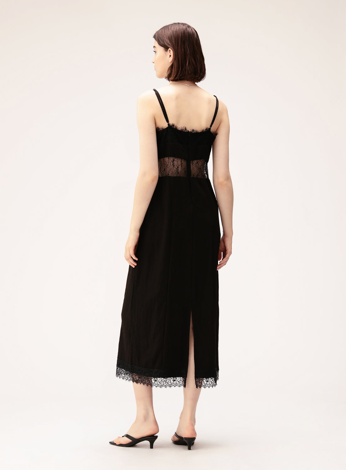 A woman stands facing away, wearing the Ray BEAMS Lace Cut-Out Cami Dress—a black, lingerie-inspired midi dress with delicate lace details, thin straps, a sheer lace midsection, and a back slit. She pairs it with black heeled sandals.