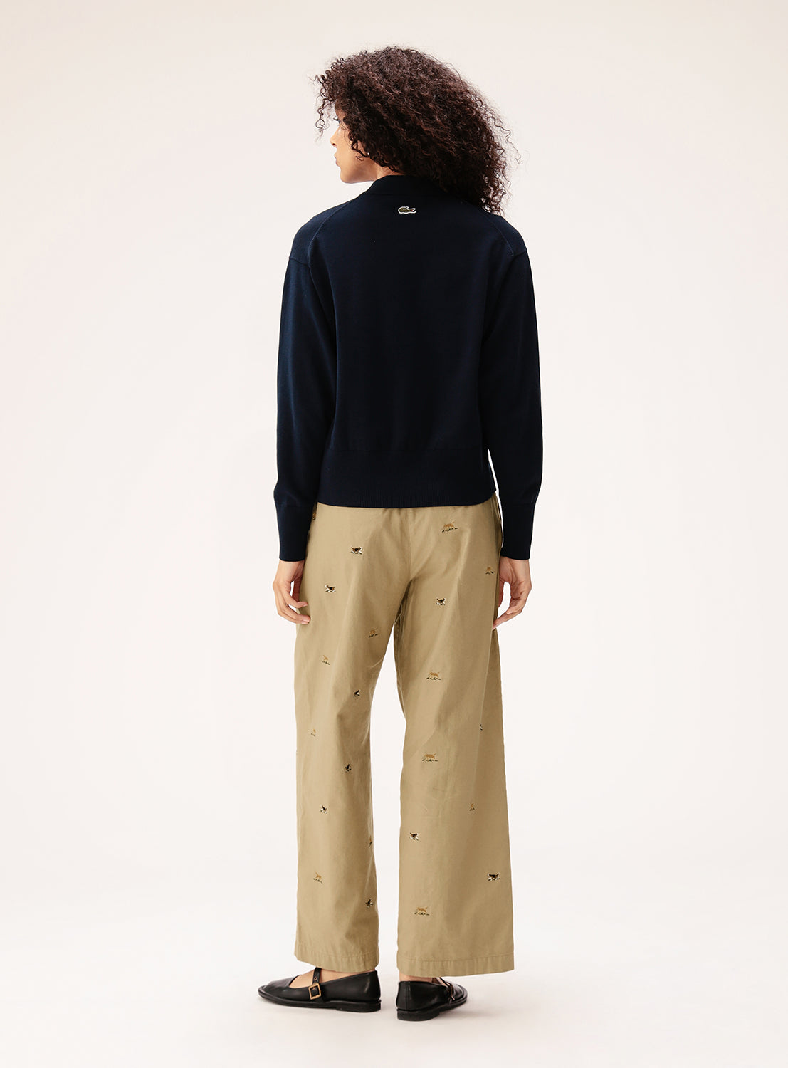 A person with curly hair stands facing away, wearing a navy sweater, BEAMS BOY Dog Embroidered Chino Pants, and black shoes against a plain light background.