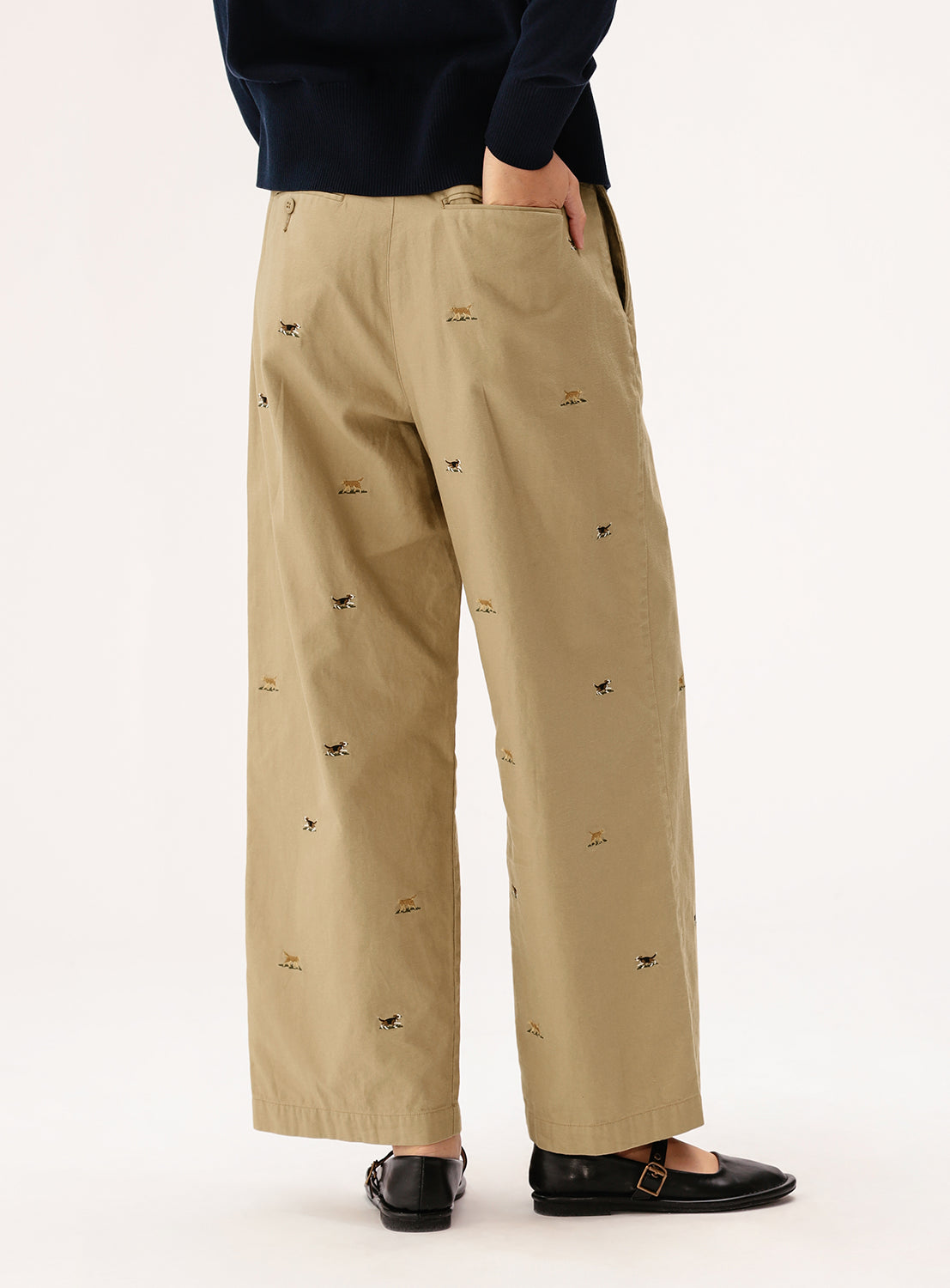 A person wears BEAMS BOY Dog Embroidered Chino Pants—loose-fitting tan chinos with small dog motifs—paired with black loafers and a partially visible dark sweater, standing with one hand in their pocket.