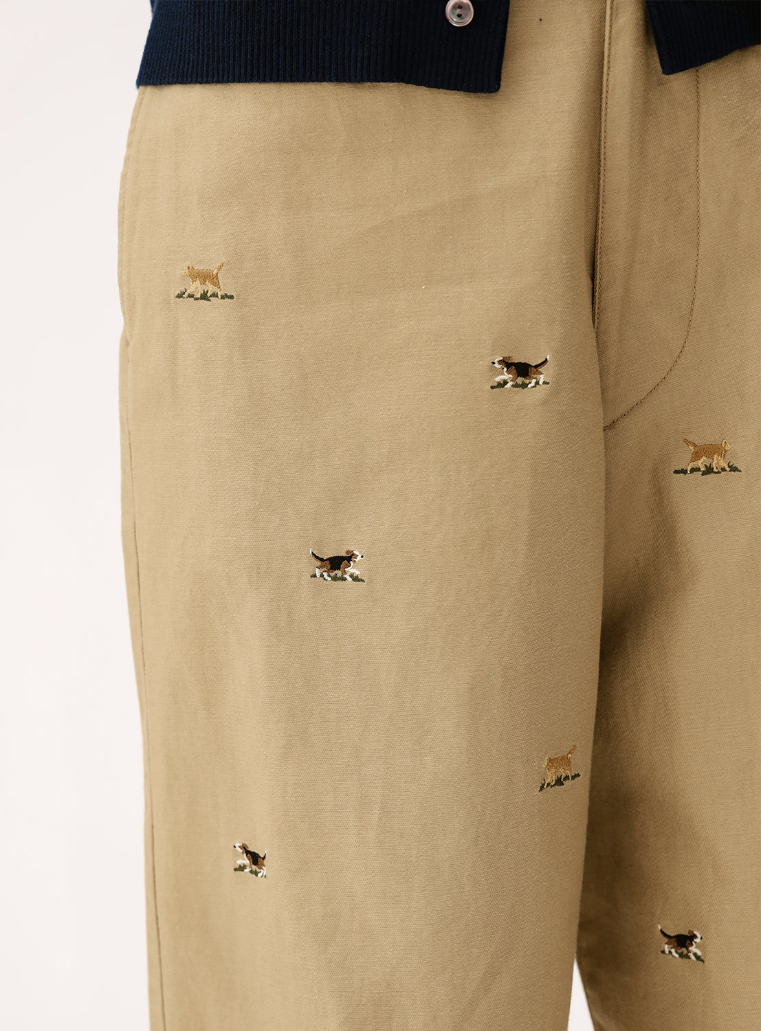 Close-up of BEAMS BOY Dog Embroidered Chino Pants, showcasing small brown, white, and black dogs stitched on tan cotton twill. The waistband and a navy garment appear at the top.