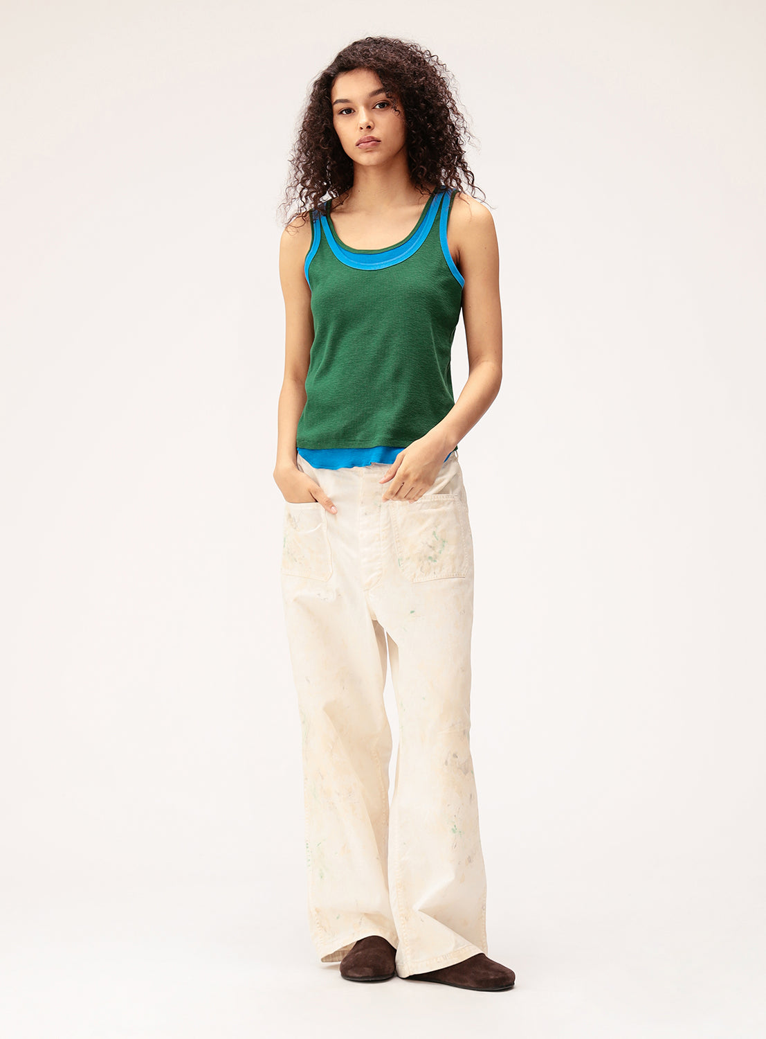 Wearing the BEAMS BOY Double-Layered Sheer Ribbed Tank Top in green with blue trim, a person with curly hair pairs it with loose white pants bearing paint stains and dark shoes. They stand against a plain, light background with their hands in their pockets.