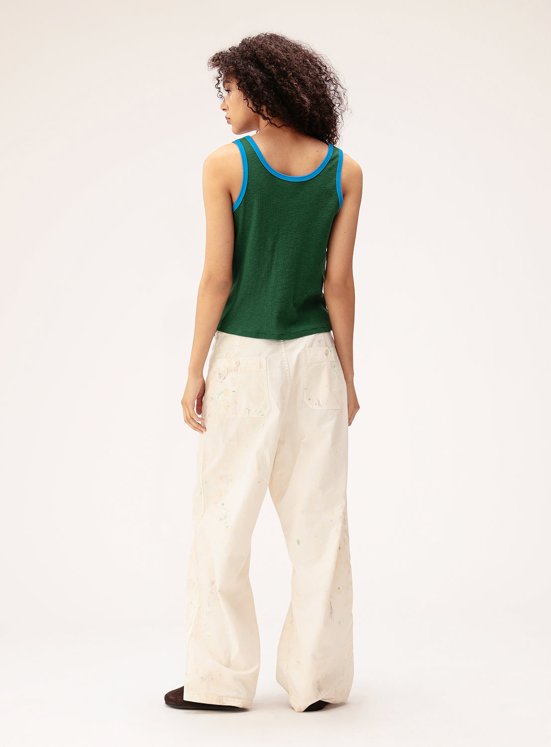 A person stands facing away, wearing the BEAMS BOY Double-Layered Sheer Ribbed Tank Top in green with blue trim, paired with loose white pants featuring paint stains. They have curly hair and are set against a plain, light-colored background.