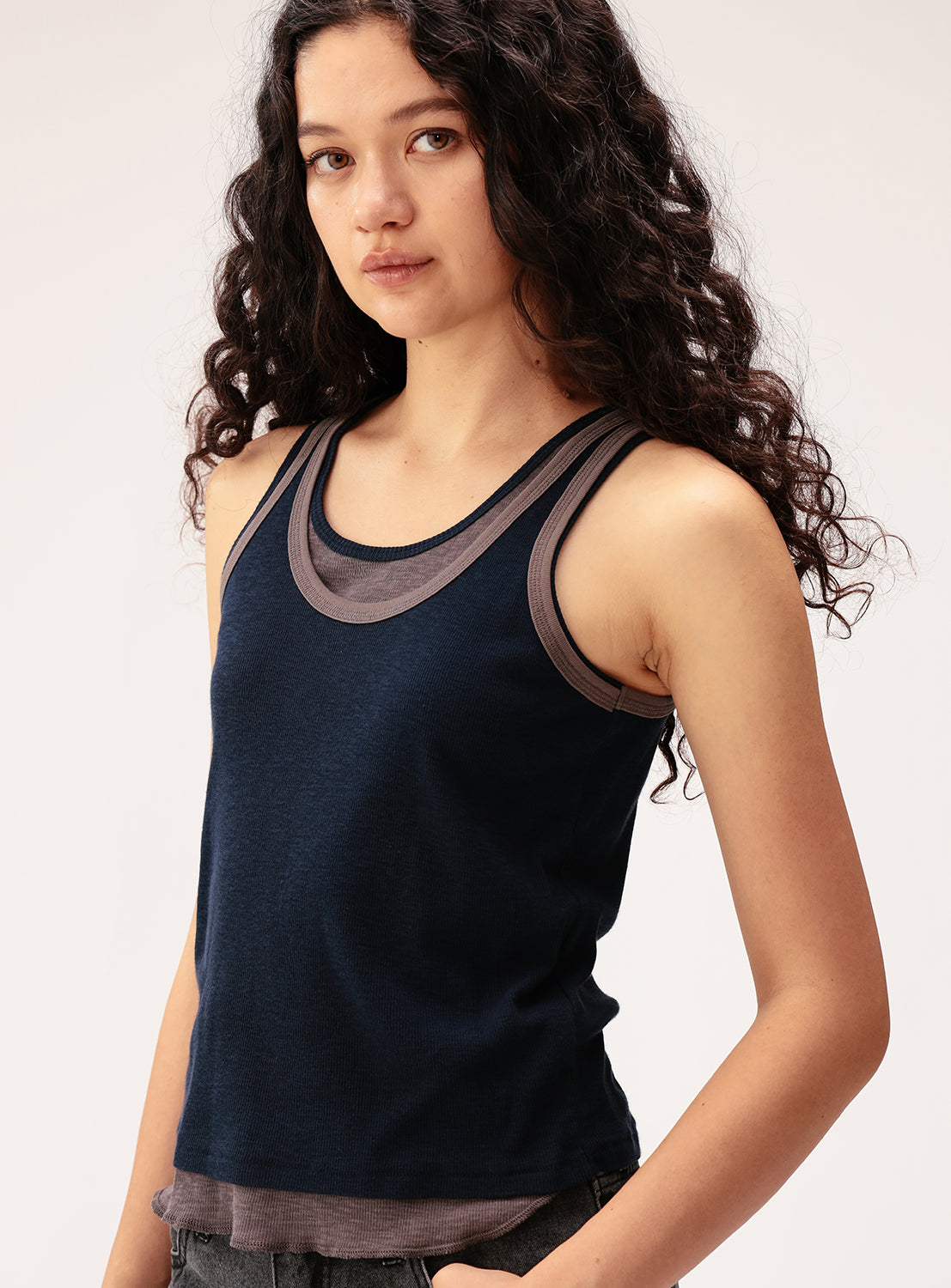 A young woman with long, curly dark hair is wearing the BEAMS BOY Layered Rib Tank Top, a navy blue cotton rib knit style featuring gray trim. She stands against a light background, gazing slightly to the side with a neutral expression.