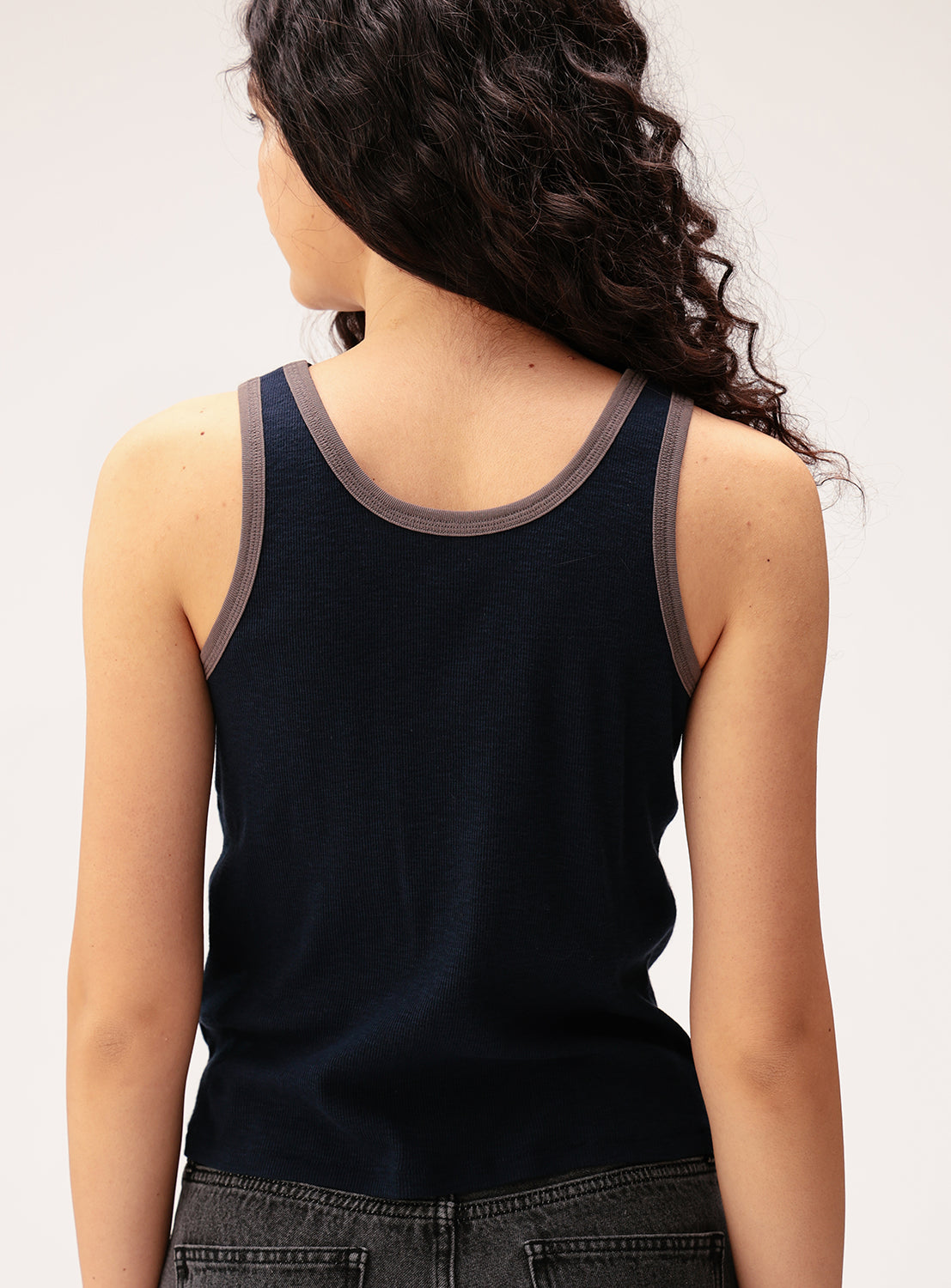 A woman with long, curly brown hair is pictured from the back, wearing the BEAMS BOY Layered Rib Tank Top in navy blue with gray trim, paired with faded black jeans. The cotton rib knit fabric of the tank adds a subtle texture to her relaxed look.