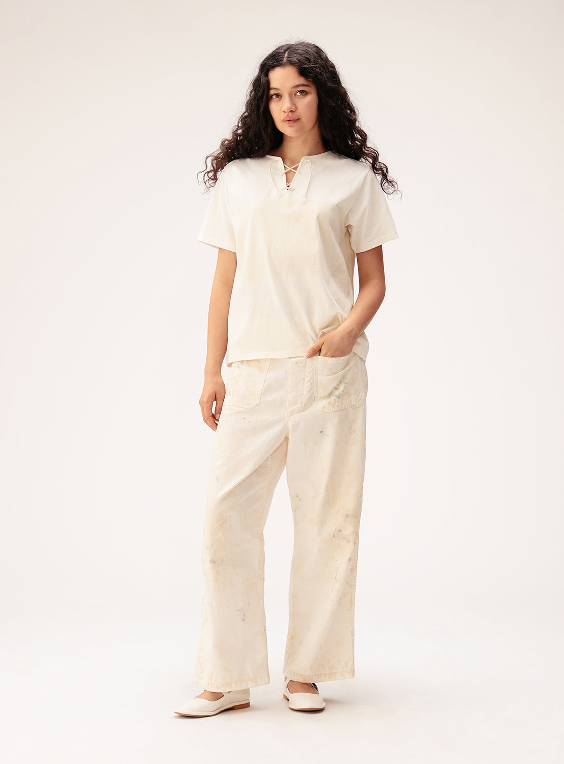 A woman with long, wavy dark hair stands against a plain background, wearing the BEAMS BOY Wide Border Lace-Up Short Sleeve Top and wide-leg light pants. One hand is in her pocket, and she completes the look with light-colored shoes.