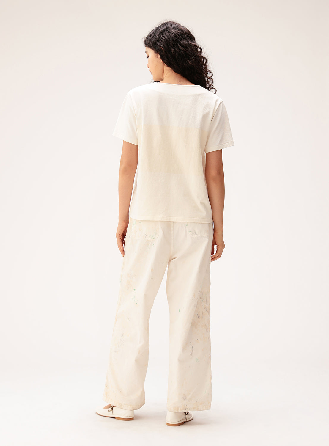 A person with long curly hair stands facing away, wearing the BEAMS BOY Wide Border Lace-Up Short Sleeve Top and loose white pants with paint stains, plus white shoes, against a plain light background.