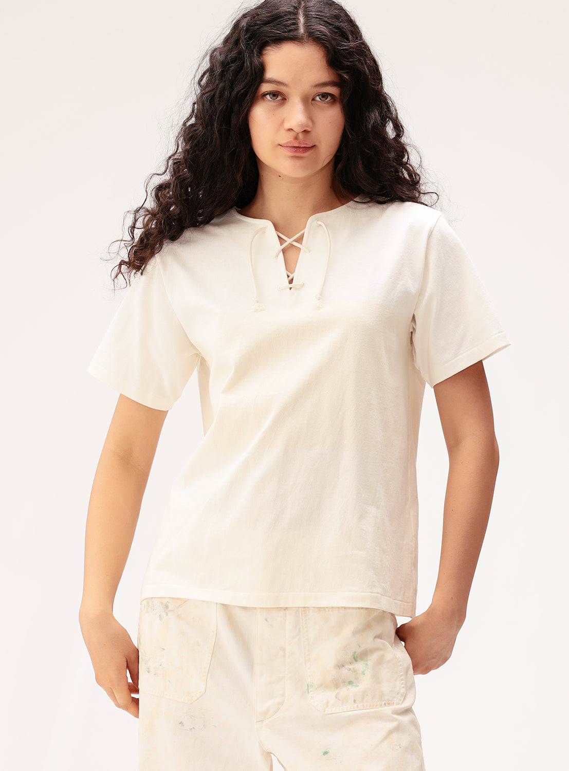 A woman with long curly hair wears the BEAMS BOY Wide Border Lace-Up Short Sleeve Top and light-colored paint-stained pants, standing against a plain white background.
