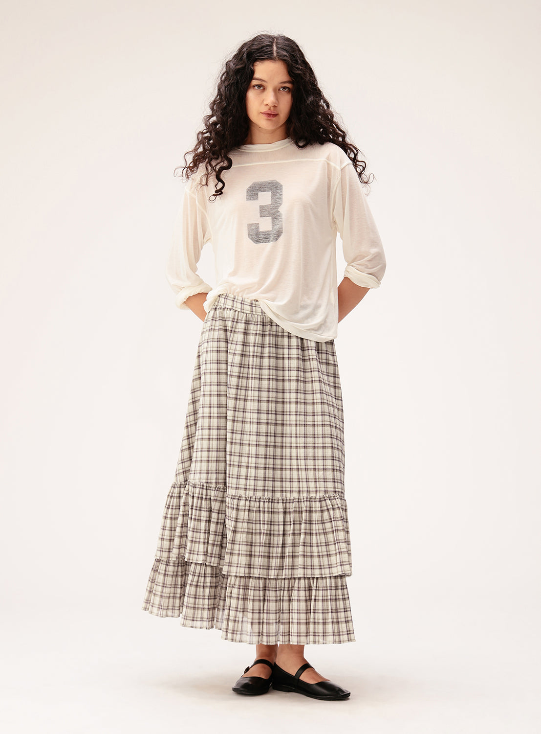 A person with long curly hair stands against a plain light background, dressed in a white shirt with the number 3, black shoes, and the BEAMS BOY Check Tiered Skirt. The skirt’s check pattern introduces a playful touch to the outfit.