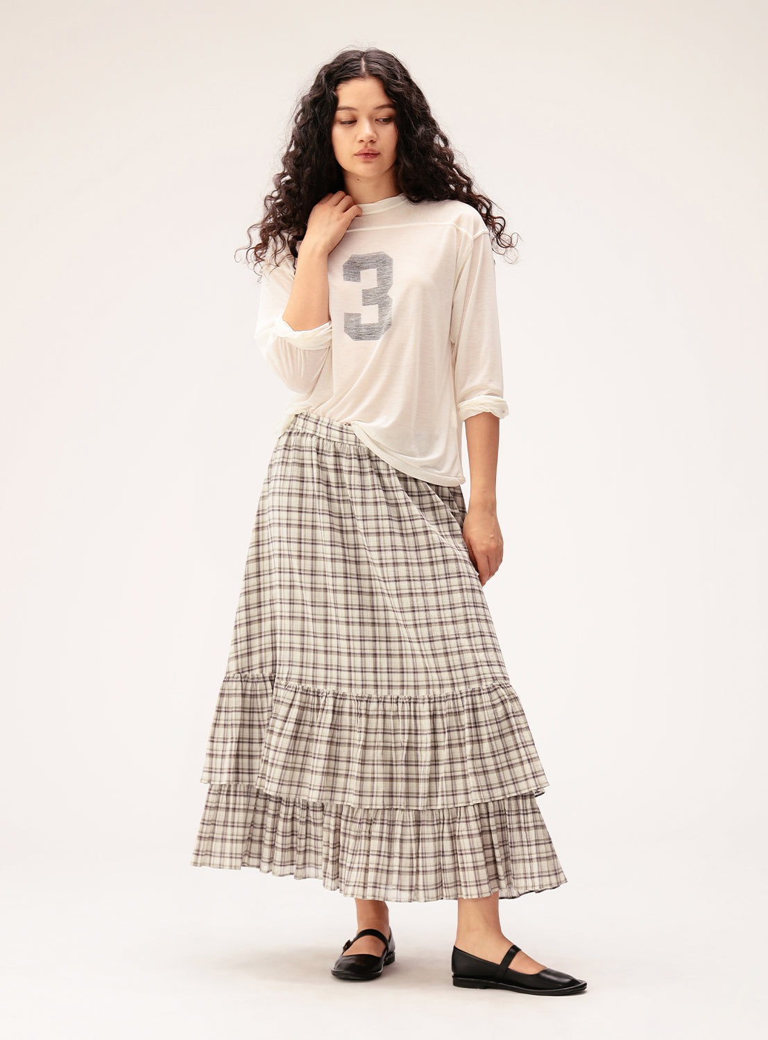 A woman with long curly hair is wearing a white top featuring a bold “3” and the BEAMS BOY Check Tiered Skirt, styled in a tiered silhouette crafted from lightweight fabric. She stands against a plain background, gazing downward with one hand gently touching her hair.