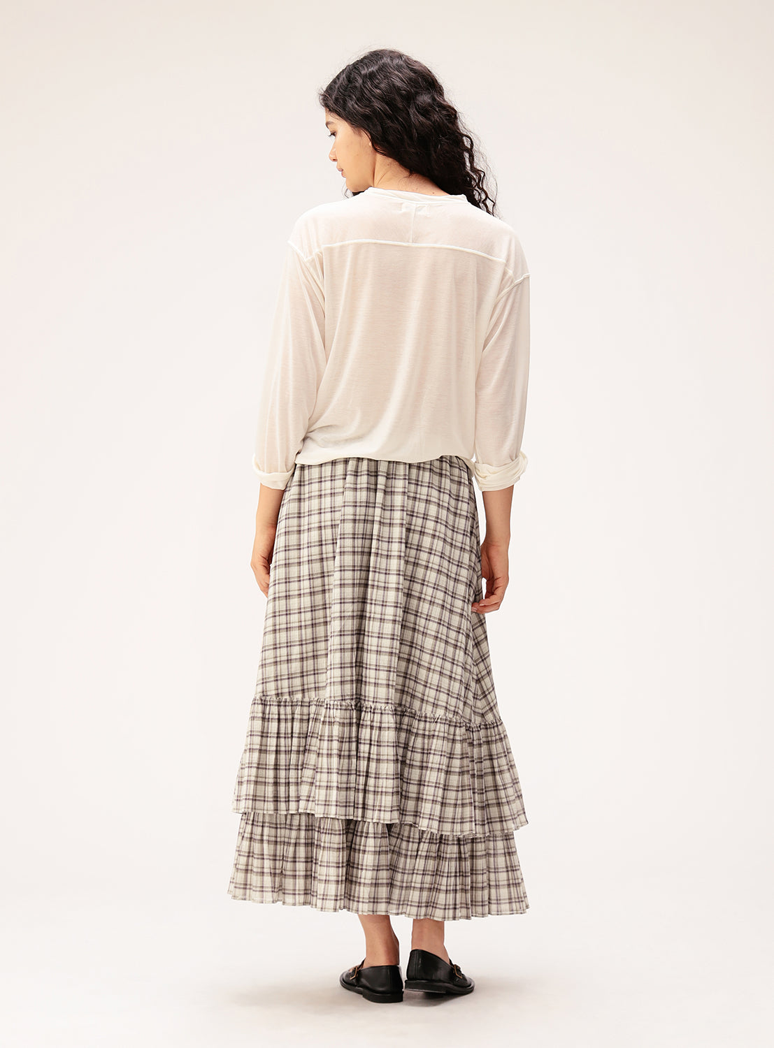 A woman, with long wavy dark hair, faces away while wearing the BEAMS BOY Check Tiered Skirt—crafted from lightweight fabric featuring the brand's signature check pattern—paired with a sheer white top and black flats against a plain light background.
