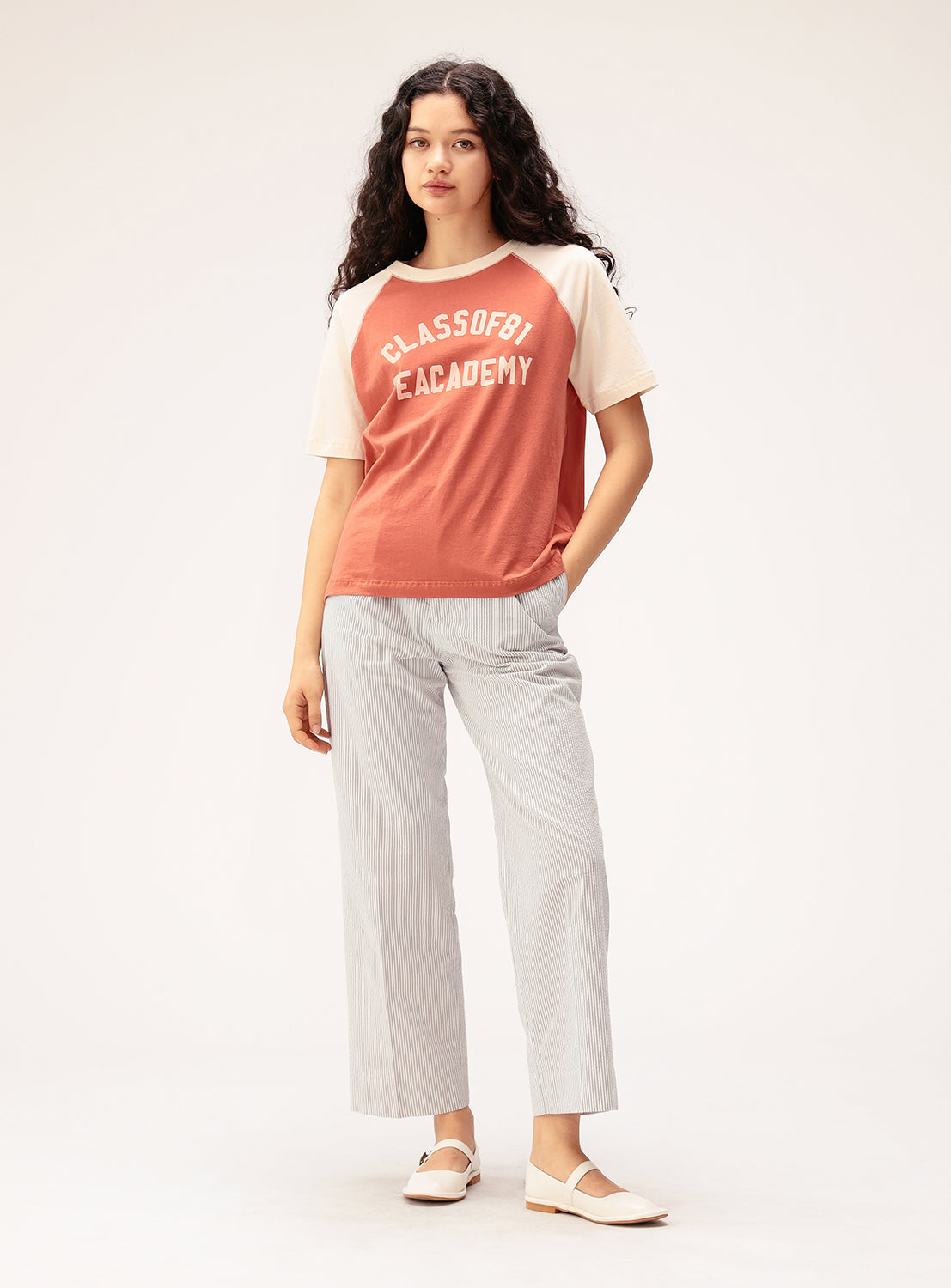 A woman with long curly hair stands against a plain light background, one hand in her pocket, exuding an effortlessly cool collegiate vibe in a BEAMS BOY Two-Tone Vintage Raglan T-shirt with a "Class of 97 Academy" print, paired with light striped pants and white shoes.