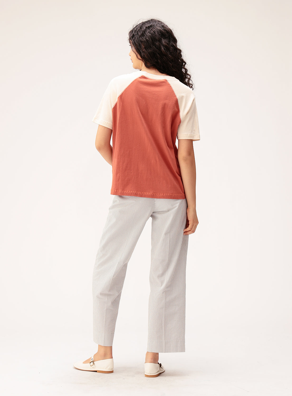 A person with long curly hair stands facing away in a studio with a plain background, wearing the BEAMS BOY Two-Tone Vintage Raglan T-shirt in red and cream, styled with light gray striped pants and beige loafers.