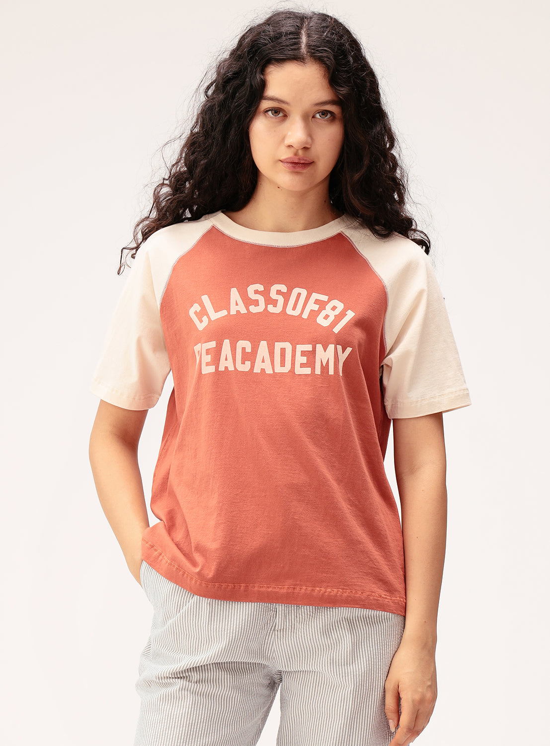 A woman with long curly hair wears the BEAMS BOY Two-Tone Vintage Raglan T-shirt in red and beige, featuring a CLASS OF 81 TEA ACADEMY graphic for a classic collegiate look. She stands against a light background with one hand in her pocket.