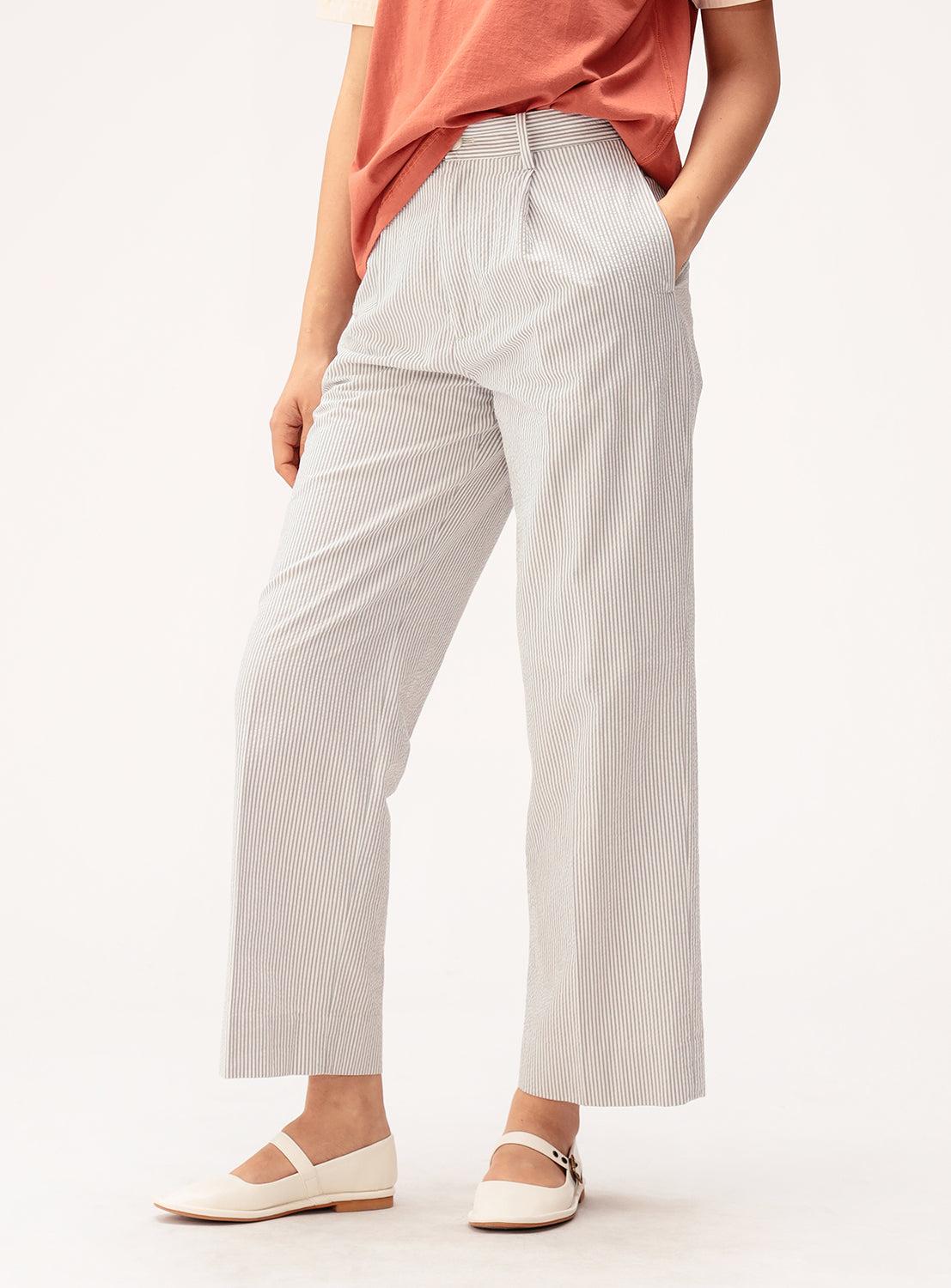 A woman stands with her hands in her pockets against a plain background, exuding effortless summer comfort in a rust-colored top, BEAMS BOY Relaxed Fit Seersucker Slacks in a light pinstripe, and white Mary Jane shoes.