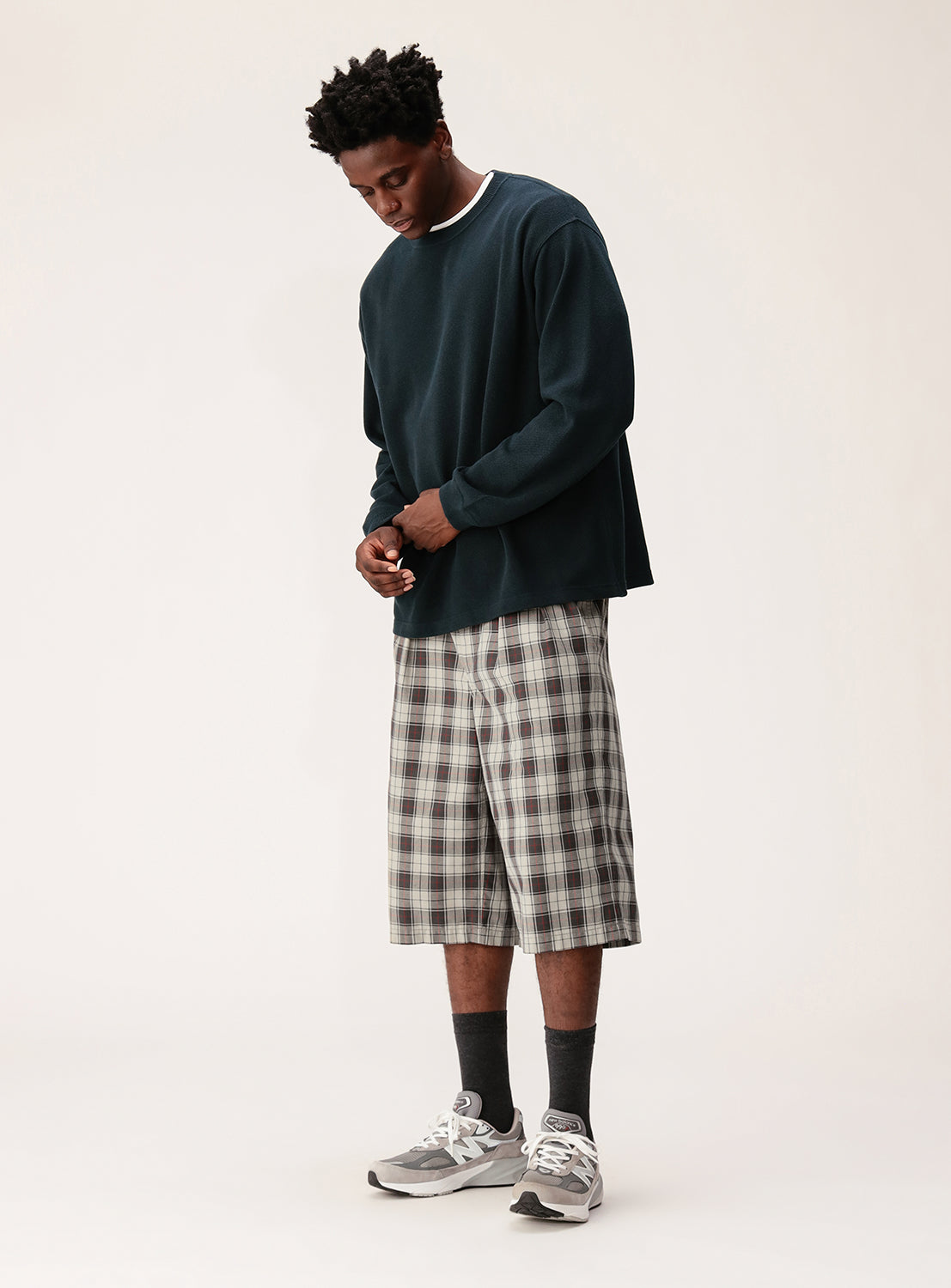 A man wearing BEAMS Tartan Check Shorts with a dark long-sleeve shirt, dark socks, and gray sneakers stands against a plain light background, looking down with his hands together.