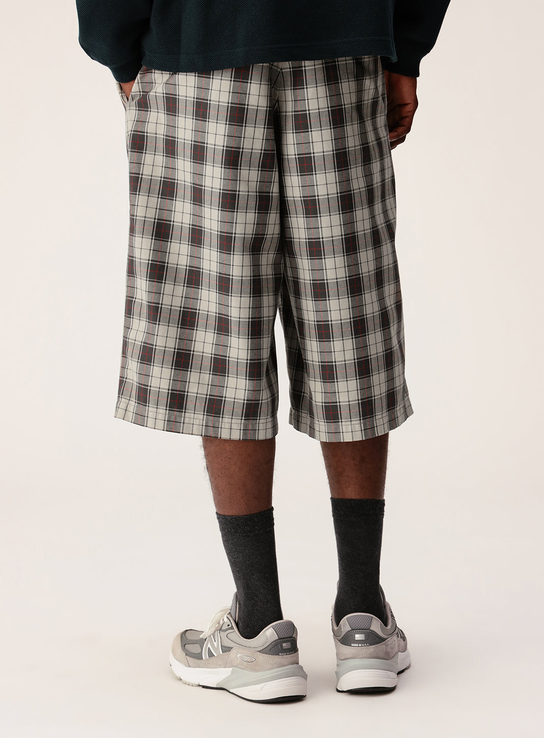 A person faces away, dressed in BEAMS Tartan Check Shorts, dark socks, gray sneakers, and a dark long-sleeve top. The background is plain and light-colored.