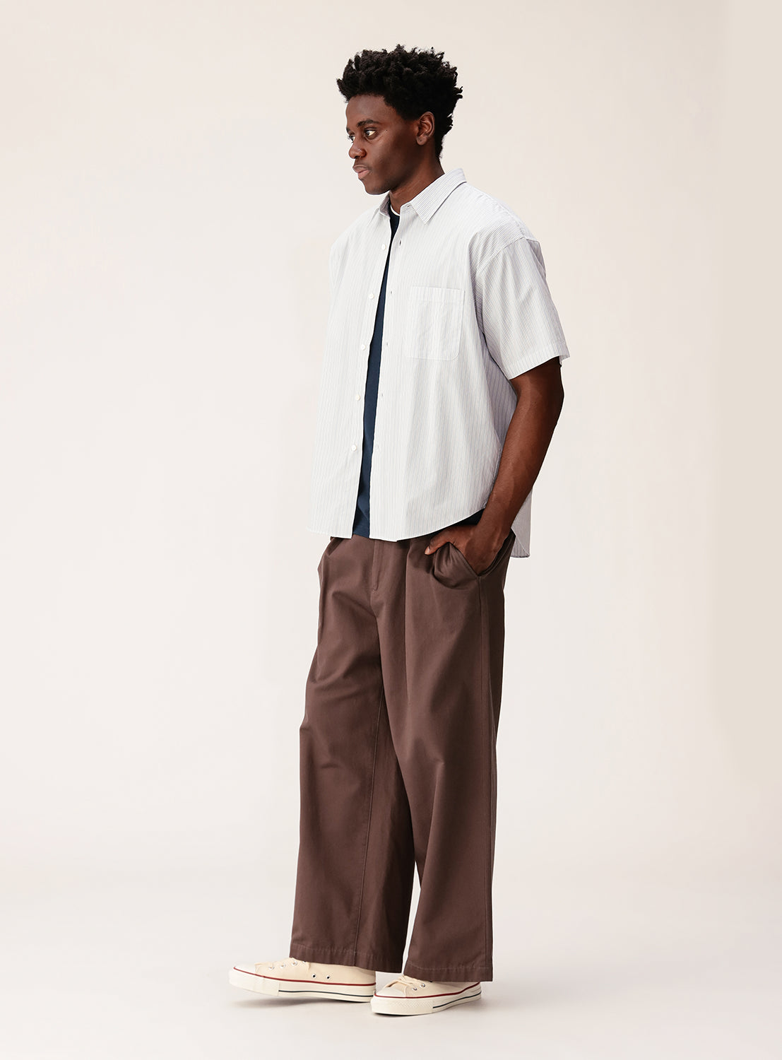 A man stands in profile wearing the BEAMS Relaxed Fit Striped Shirt featuring light, broad stripes and a mini-regular collar, paired with wide brown pants and cream sneakers against a plain light background.