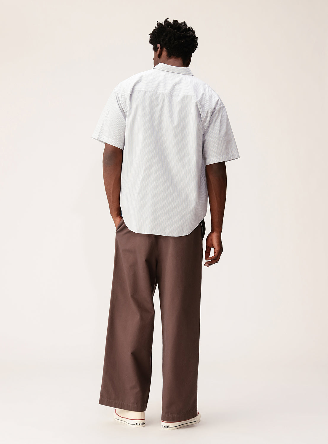 A man stands facing away, wearing the BEAMS Relaxed Fit Striped Shirt—a light blue, short-sleeve button-up—paired with wide brown pants and white sneakers against a plain, light background.