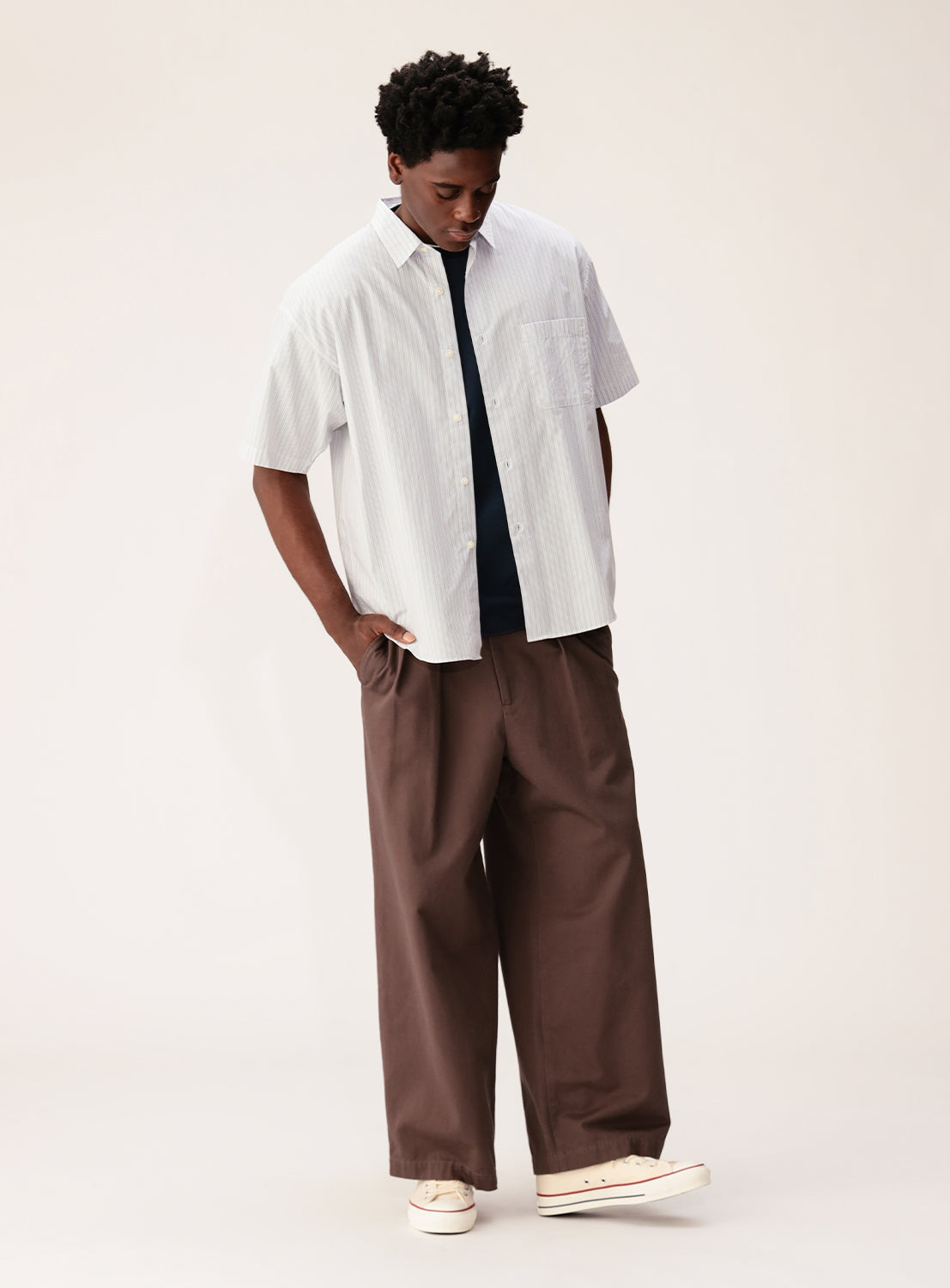 A person wearing the BEAMS Relaxed Fit Striped Shirt with short sleeves over a dark T-shirt, paired with wide brown pants and cream sneakers, stands against a plain light background, looking down with hands in pockets.