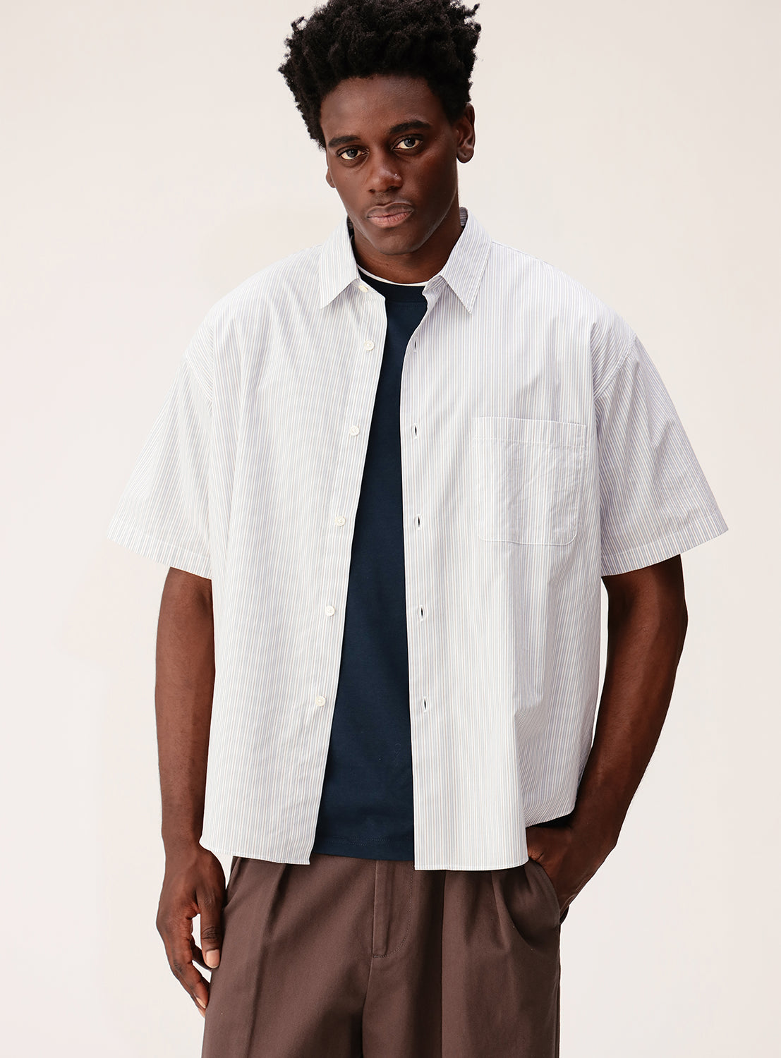 A man wears the BEAMS Relaxed Fit Striped Shirt in light blue, styled over a dark t-shirt and paired with brown pants, standing against a plain light background.