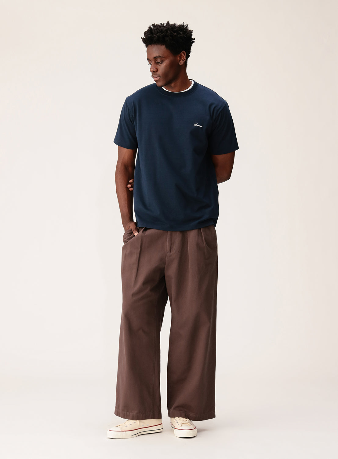 A man models the BEAMS Signature Logo Basic Fit T-Shirt in navy, paired with loose brown pants and white sneakers. He stands against a plain light background, gazing left with his hands in his pockets, embodying a minimalist design aesthetic.
