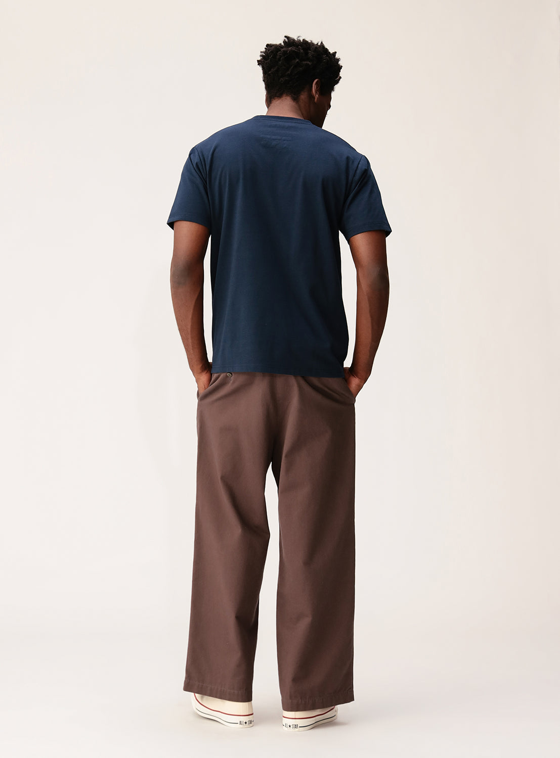 A person stands facing away, wearing the BEAMS Signature Logo Basic Fit T-Shirt in navy blue with a subtle minimalist design, paired with loose brown pants and white shoes, against a plain light background.