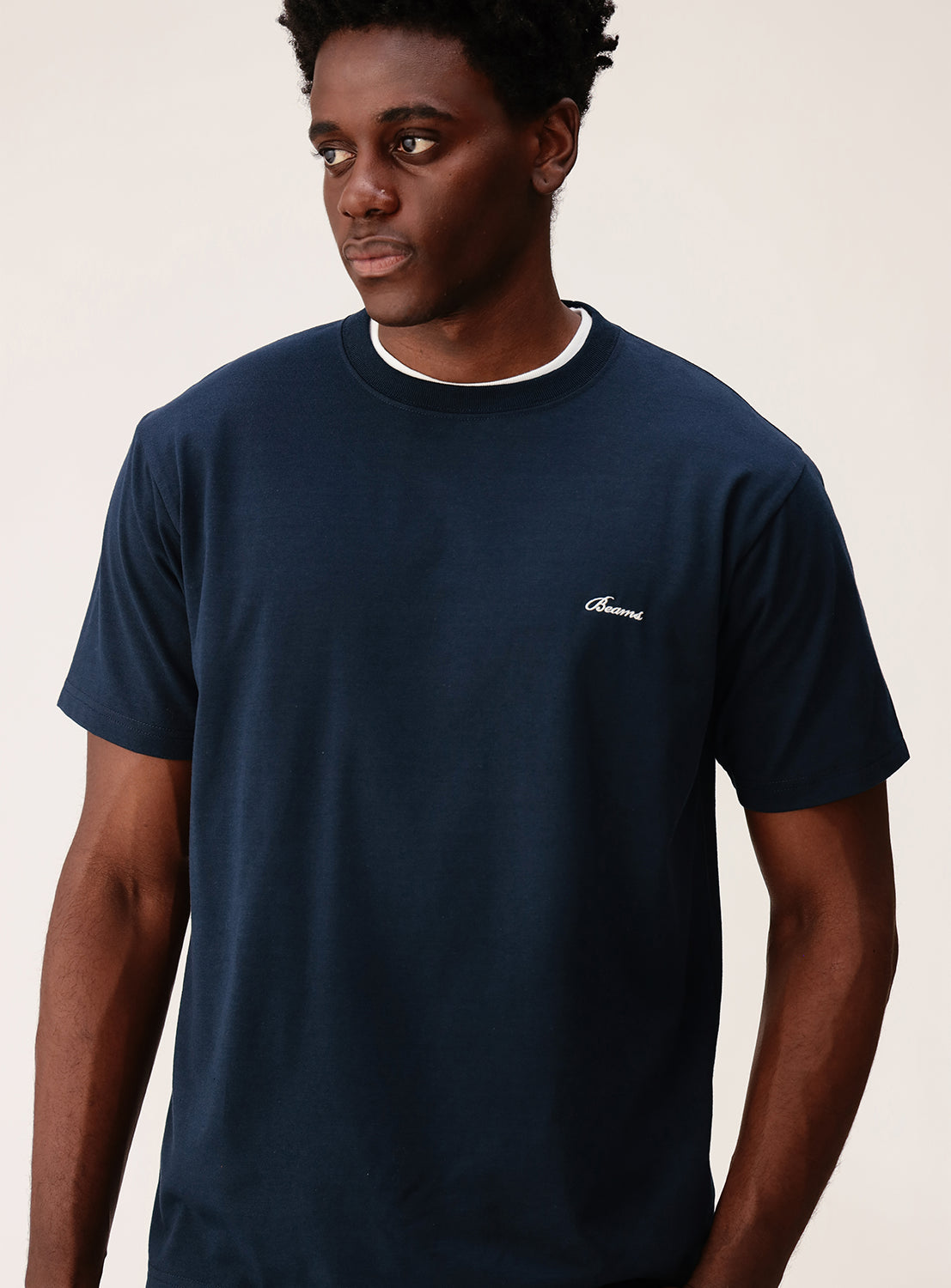 A man stands against a plain light background, looking slightly to his left, wearing the BEAMS Signature Logo Basic Fit T-Shirt in navy blue. The crew neck T-shirt features a small white BEAMS logo on the left chest, and its minimalist design enhances the clean and modern aesthetic.