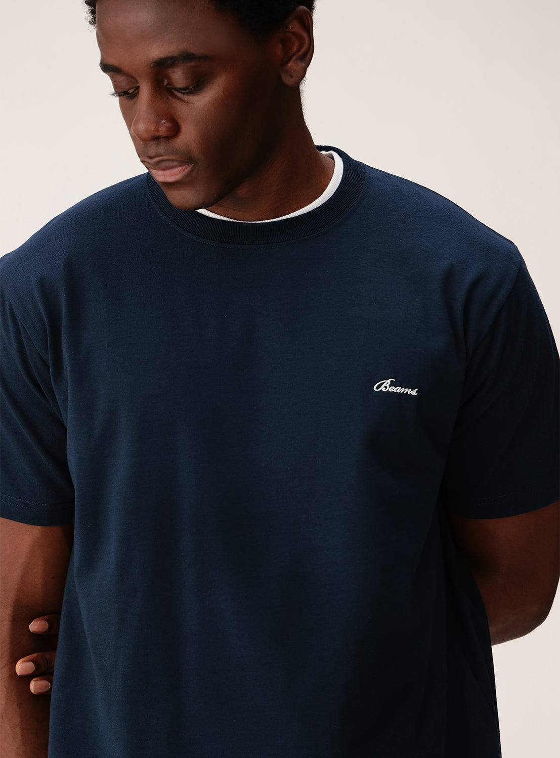A person wearing the BEAMS Signature Logo Basic Fit T-Shirt in dark blue, featuring the brand's logo embroidered on the chest, looks down with a neutral expression against a plain background, highlighting its minimalist design.