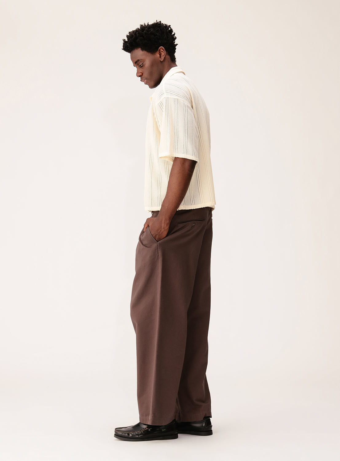 A man stands in profile against a plain background, wearing a BEAMS Mesh Open Collar Short Sleeve Shirt, loose brown trousers, and black shoes. His hands are in his pockets as he looks down.