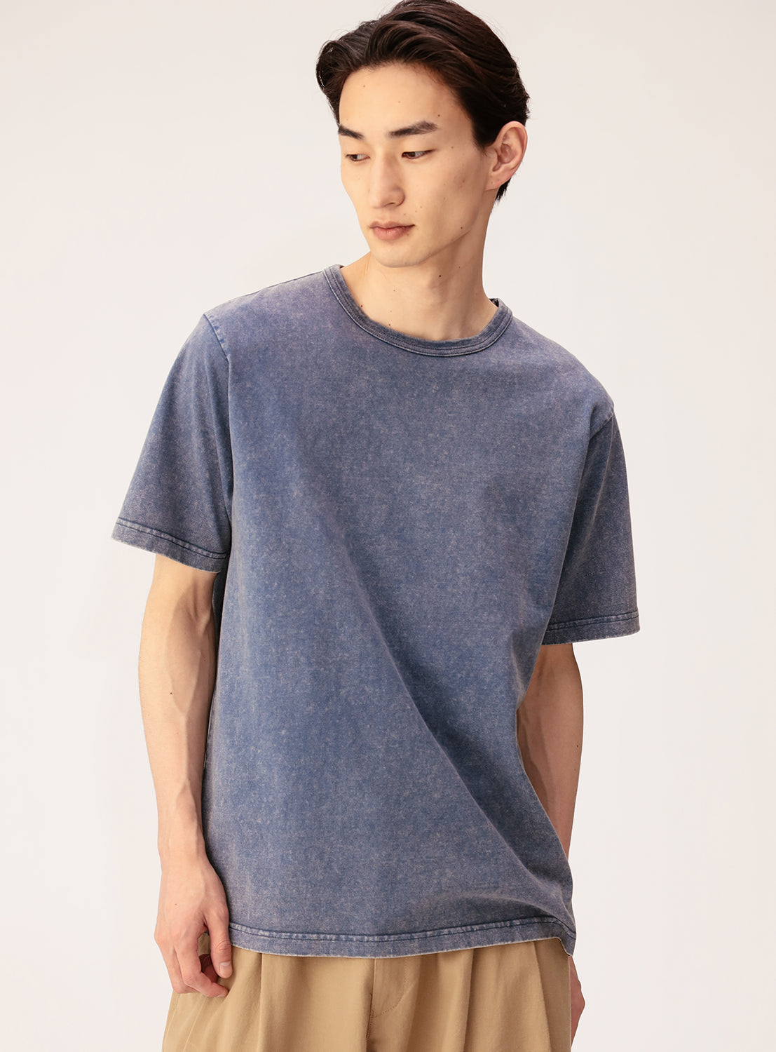 A young man with short dark hair wears the BEAMS Ultra Faded Crew Neck T-Shirt in midweight cotton jersey paired with beige pants, standing against a plain light background and looking down to his left with a neutral expression.