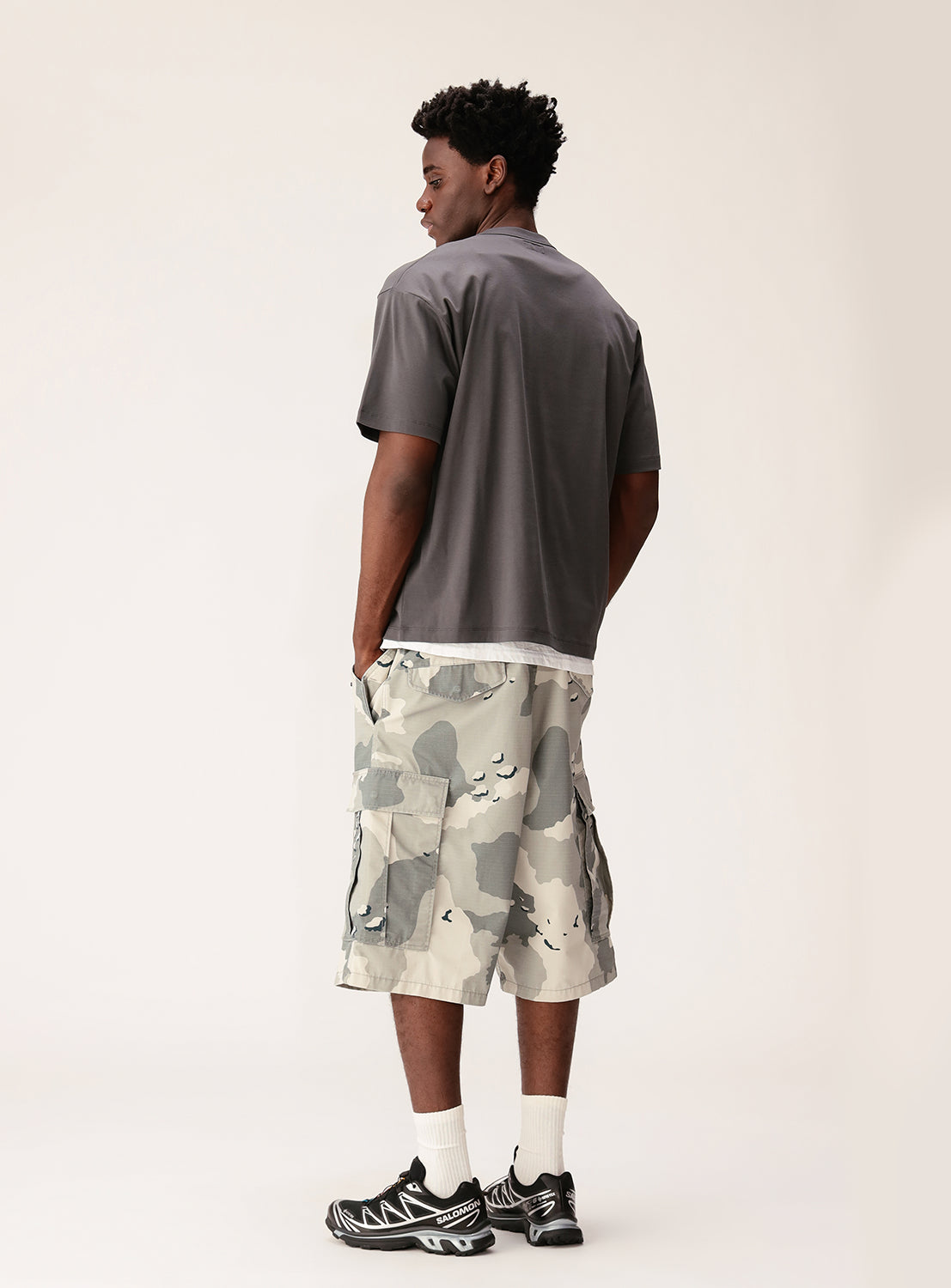 A man stands facing away against a plain light background, wearing the BEAMS Silky Relaxed Fit Crew Neck T-Shirt in gray, layered over a white shirt, paired with camo cargo shorts and black athletic sneakers.
