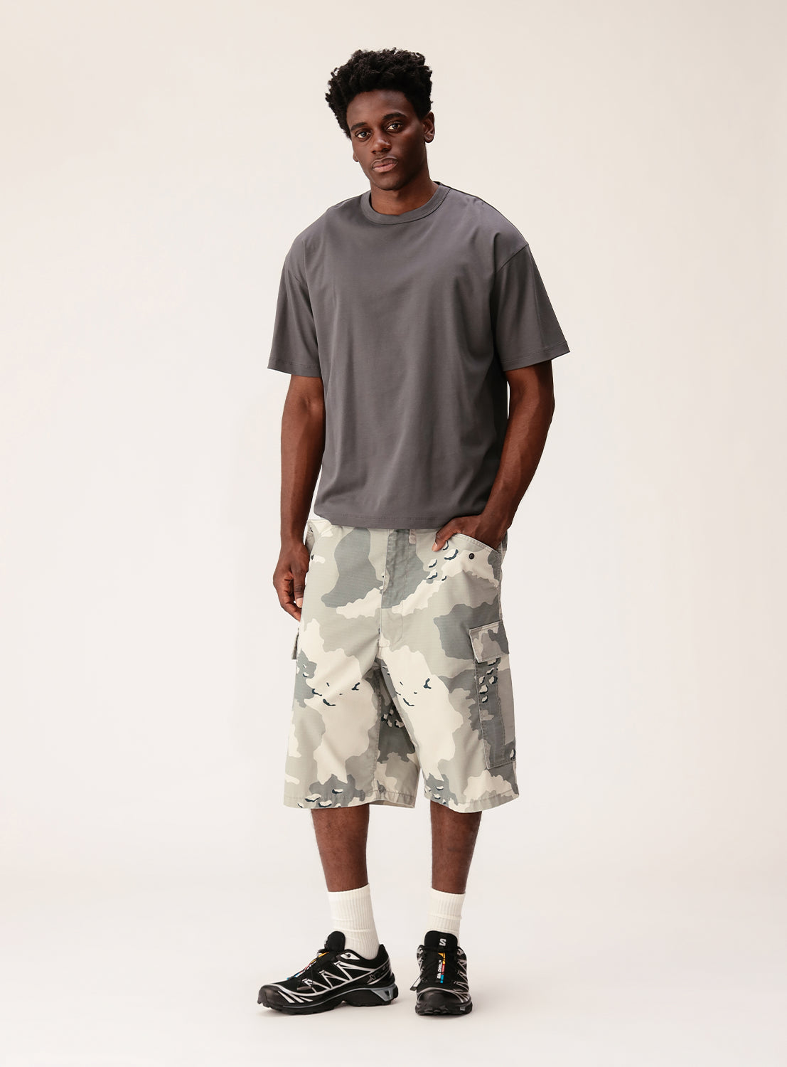 A man stands against a plain background wearing the BEAMS Silky Relaxed Fit Crew Neck T-Shirt in gray, paired with camo-patterned knee-length shorts, white socks, and black sneakers. One hand is in his pocket as he faces the camera with a neutral expression.