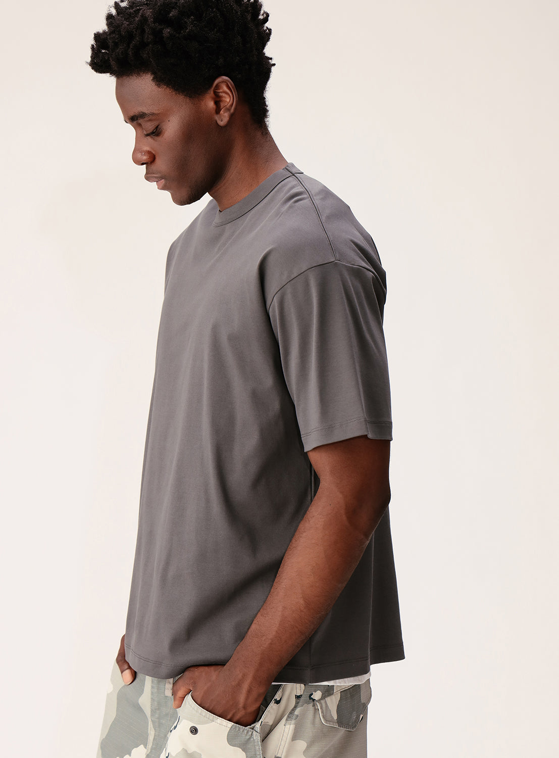 A man in a loose Silky Relaxed Fit Crew Neck T-Shirt by BEAMS and light camouflage shorts stands in profile against a plain light background, looking down with his left hand in his pocket.