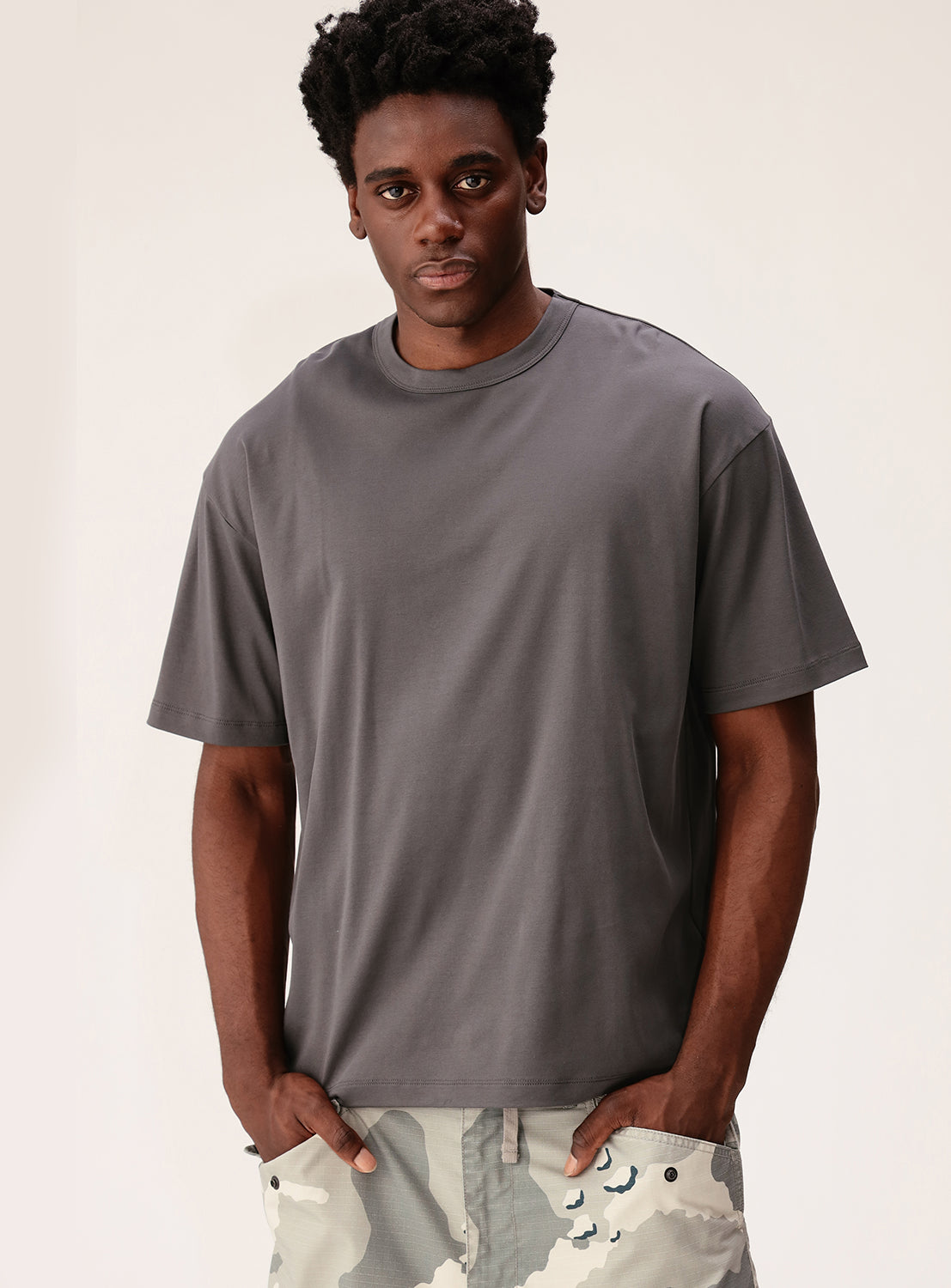 A man wearing the BEAMS Silky Relaxed Fit Crew Neck T-Shirt in gray, paired with light camouflage pants, stands against a plain light background, looking at the camera with his hands in his pockets.