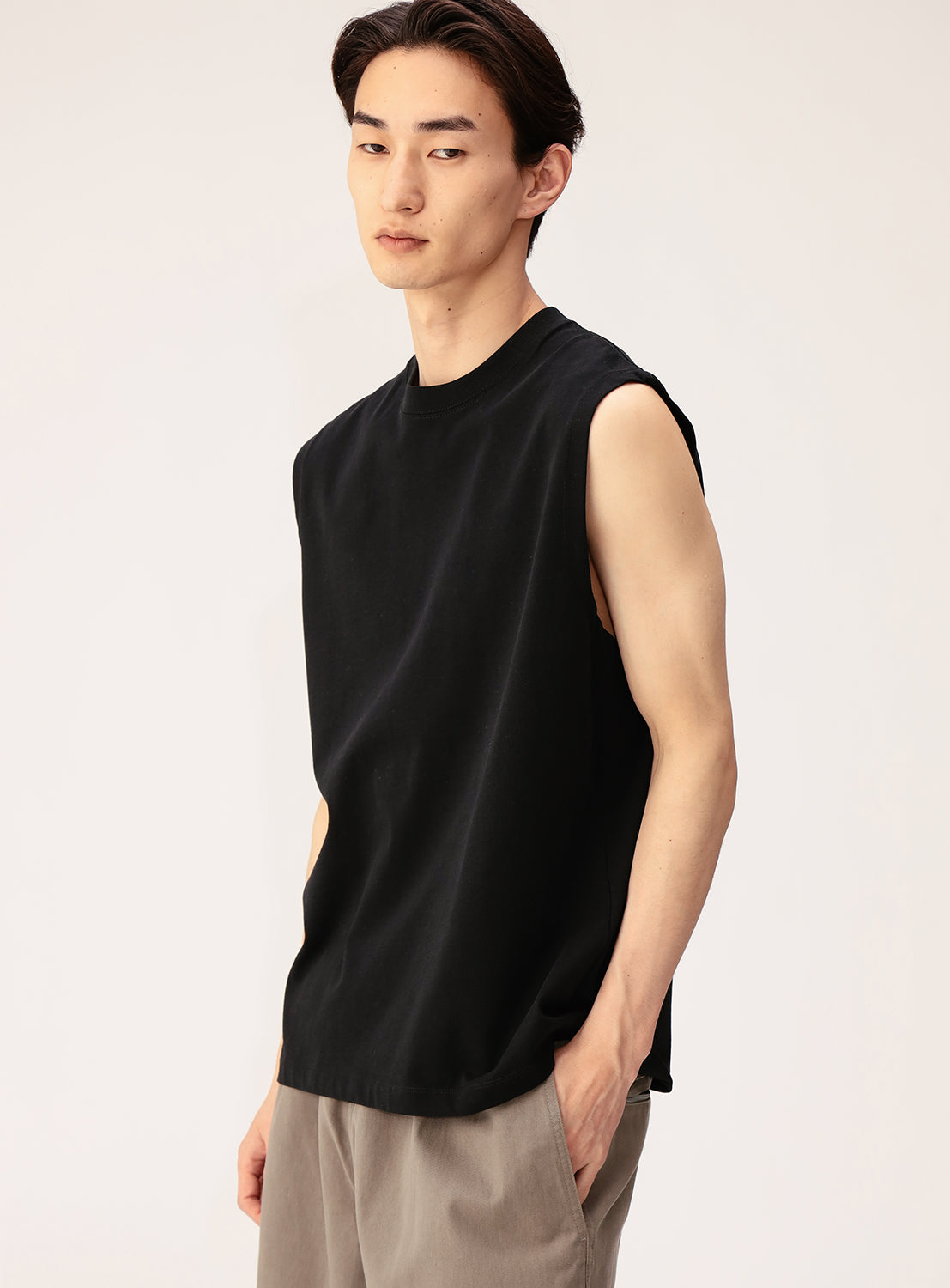 A young man with short dark hair wears a BEAMS Sleeveless Tank Top and light beige pants. The tank's relaxed fit and breathable cotton material provide comfort as he stands against a plain light background, gazing slightly to the side.