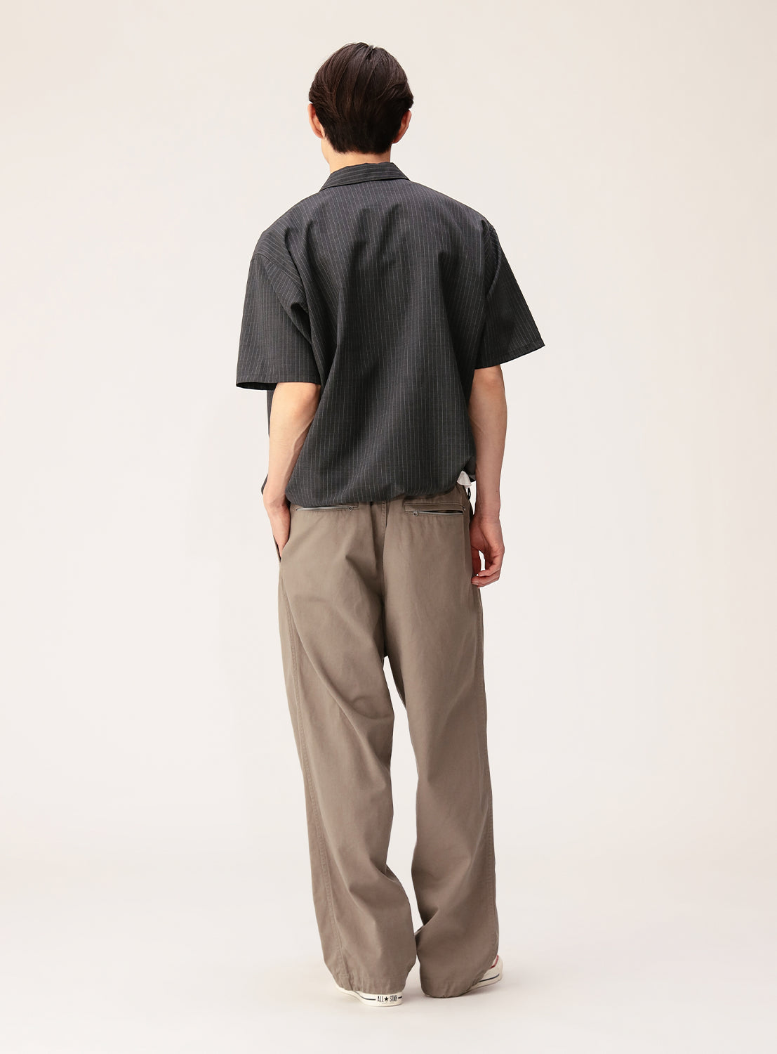 A person stands facing away, wearing the BEAMS TW Wide Fit Half-Zip Shirt in a dark pinstripe design paired with loose, light brown pants. The relaxed silhouette is complemented by the plain, light-colored background.