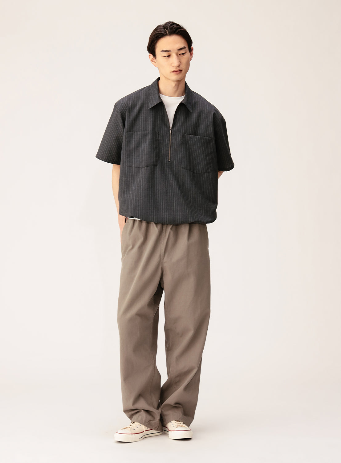 A person stands against a plain background, wearing the BEAMS TW Wide Fit Half-Zip Shirt— a short-sleeved pullover crafted from polyester-wool tropical fabric in a dark shade. The relaxed silhouette is paired with loose-fitting khaki pants and white sneakers, hands behind their back and gaze angled slightly downward.
