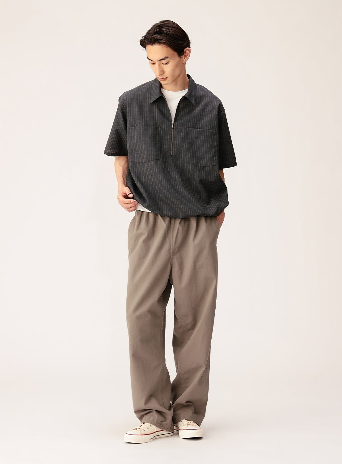 A person stands against a plain background, wearing the BEAMS TW Wide Fit Half-Zip Shirt paired with loose light brown polyester-wool tropical pants and white sneakers, looking down with one hand in their pocket.