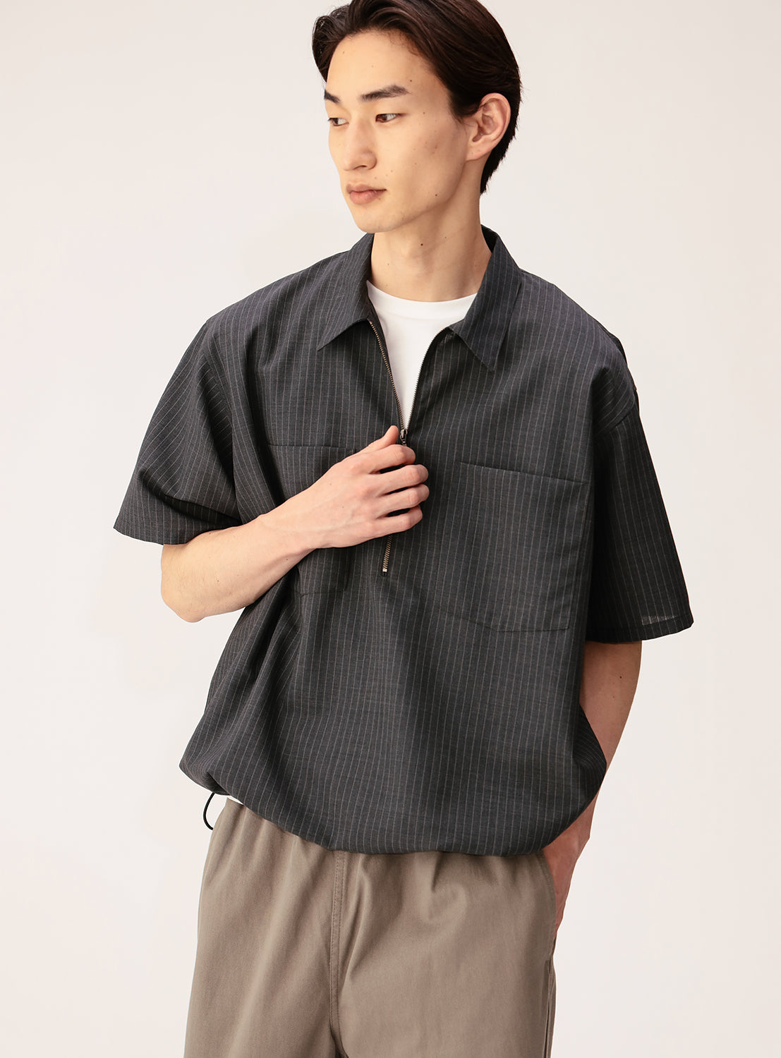 A person wearing the BEAMS TW Wide Fit Half-Zip Shirt—a short-sleeved, dark pinstripe pullover crafted from a polyester-wool tropical fabric—styled over a white tee and paired with light brown pants, stands with one hand in their pocket and looks to the side, embodying a relaxed silhouette.