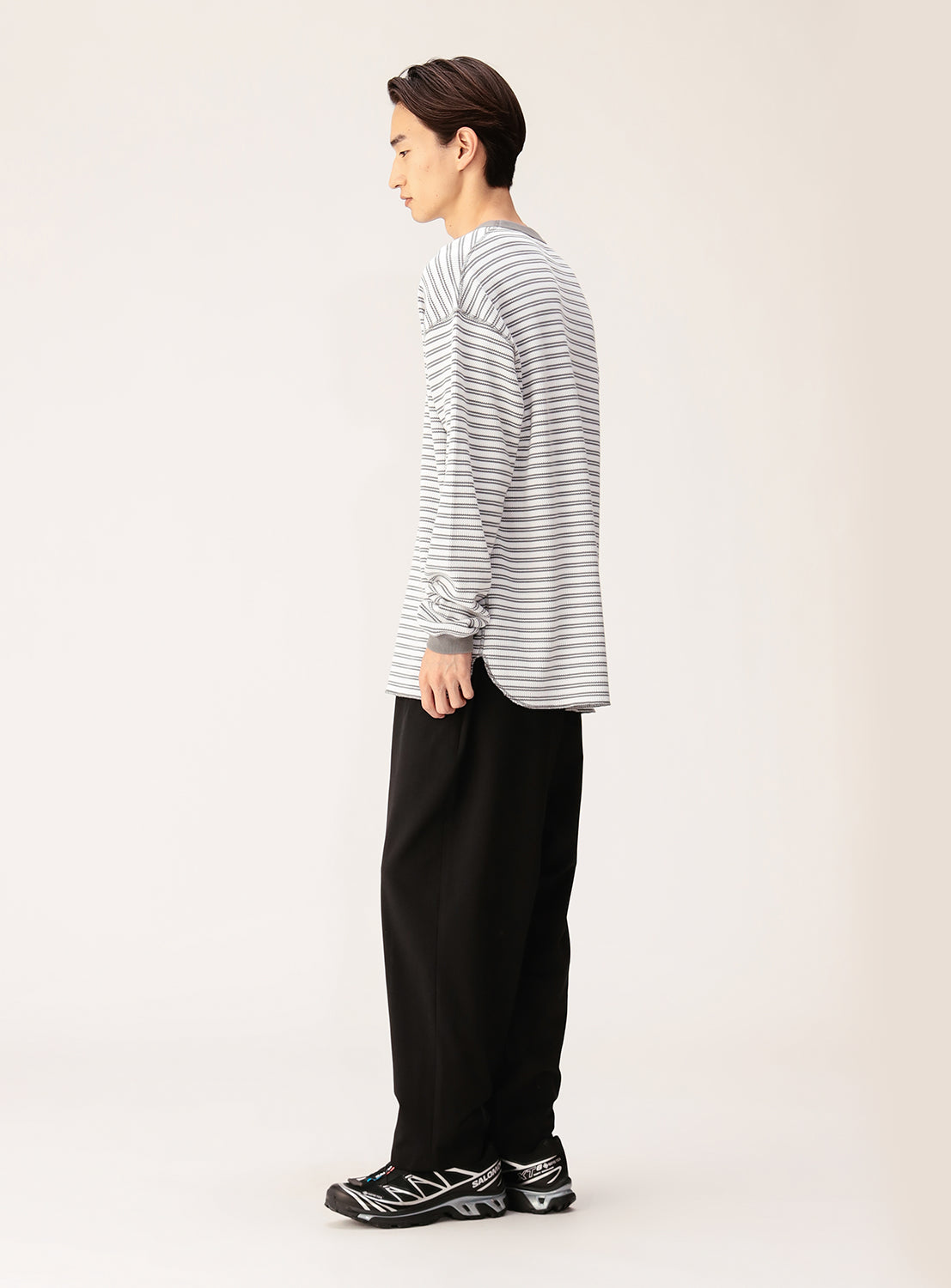 A person stands sideways against a plain light background, wearing a gray and white striped long-sleeve shirt, BEAMS Quick-Dry 2-Pleat Wide Tapered Easy Pants in black, and black sneakers with white accents.