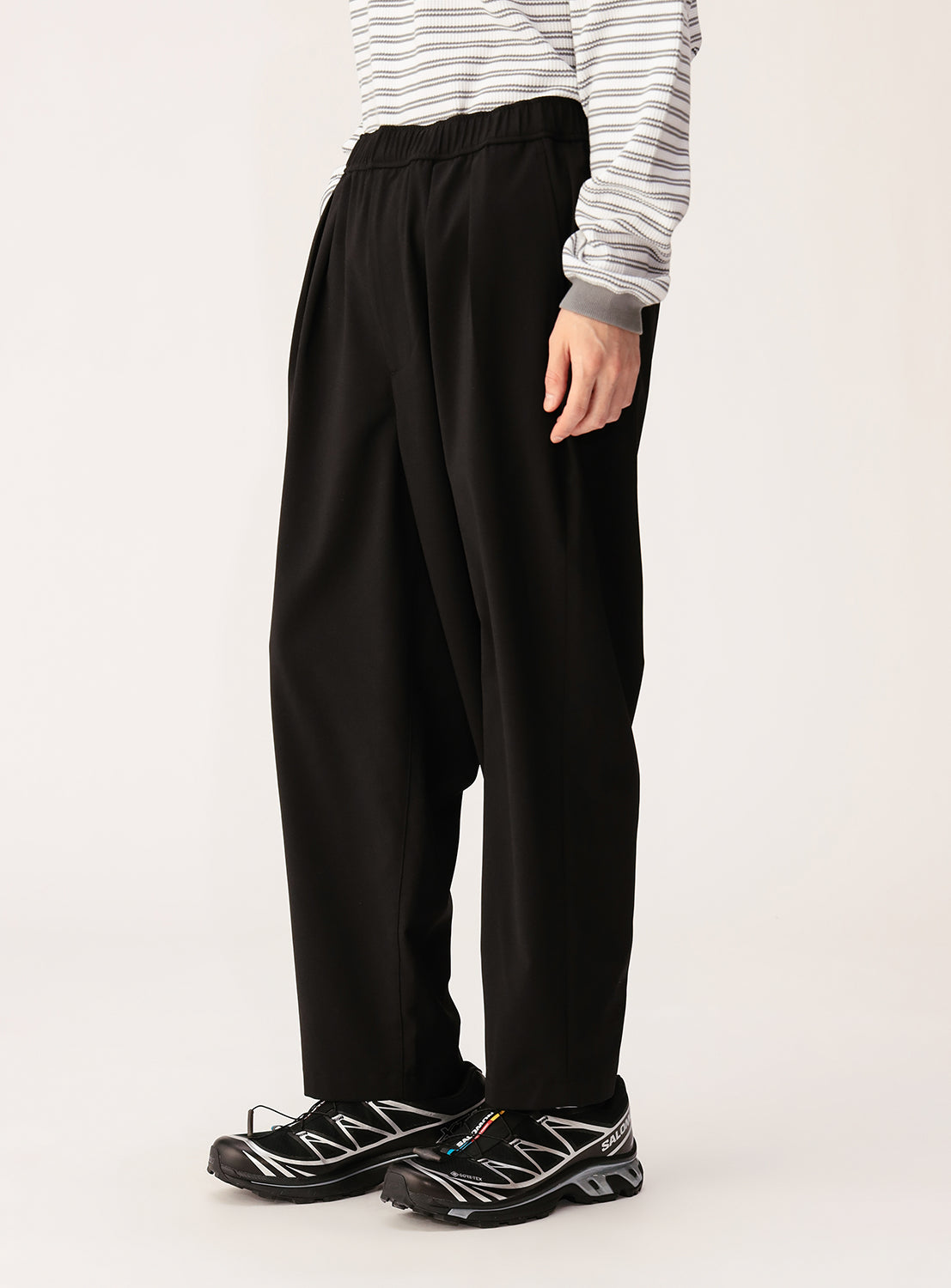 A person wears BEAMS Quick-Dry 2-Pleat Wide Tapered Easy Pants, a light striped shirt, and black sneakers with silver details, standing against a plain background. Only the lower half of their body is visible.