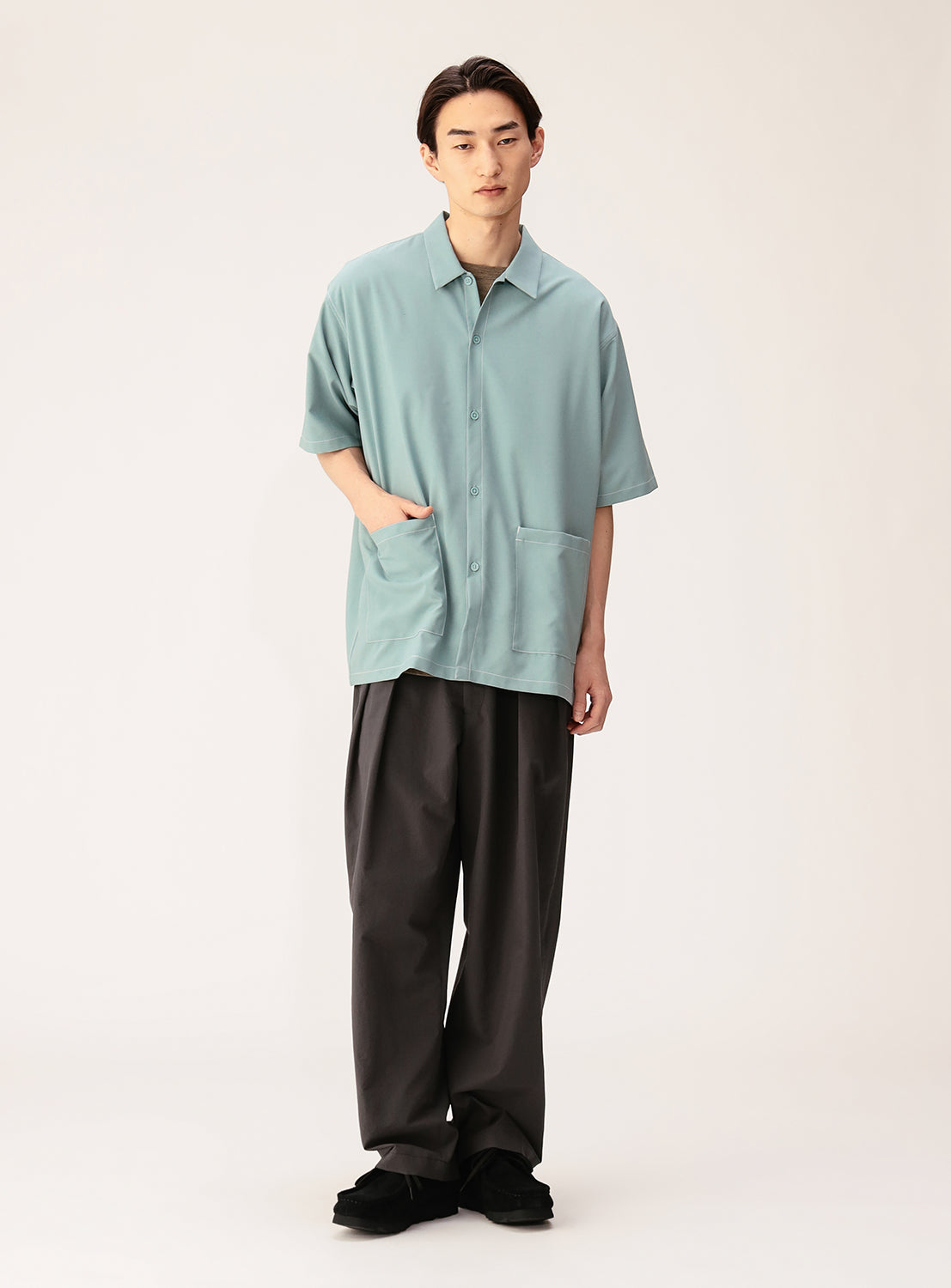 A person stands against a plain background, wearing the BEAMS Wide Fit Double Pocket Contrast Stitch Shirt in light blue, paired with wide dark pants and black shoes.