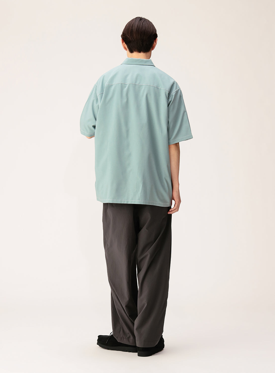 Person standing with their back to the camera, wearing the BEAMS Wide Fit Double Pocket Contrast Stitch Shirt in light blue, paired with loose dark gray pants and black shoes, against a plain light background.