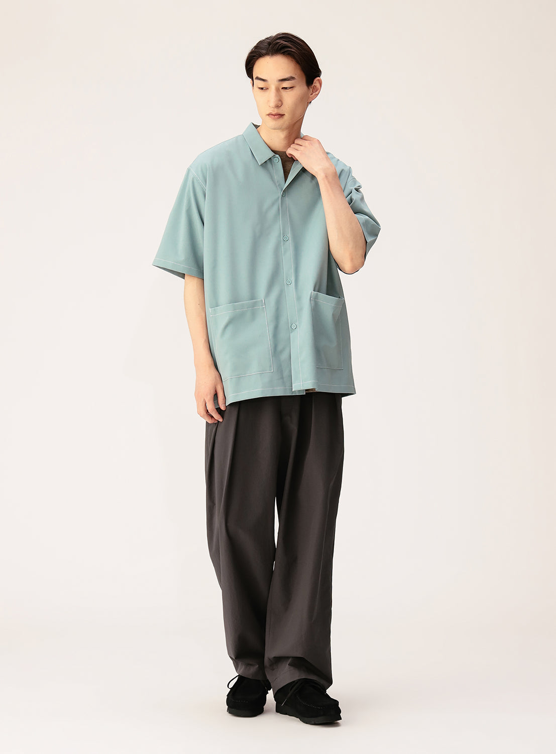 A person stands against a plain background, wearing the BEAMS Wide Fit Double Pocket Contrast Stitch Shirt—a light blue short-sleeved utility top with large front pockets and performance fabric—paired with wide dark pants and black shoes, looking slightly down with one hand touching their neck.