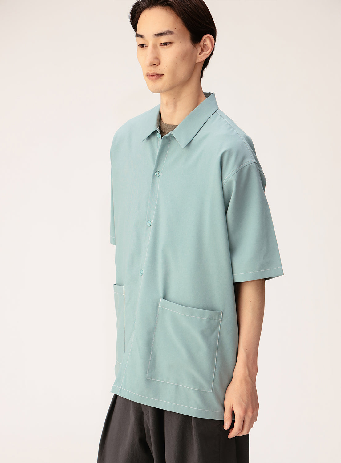 A person with straight dark hair wears the BEAMS Wide Fit Double Pocket Contrast Stitch Shirt—a loose, light blue short-sleeve utility shirt featuring two large front pockets—paired with dark pants, standing against a plain light background.